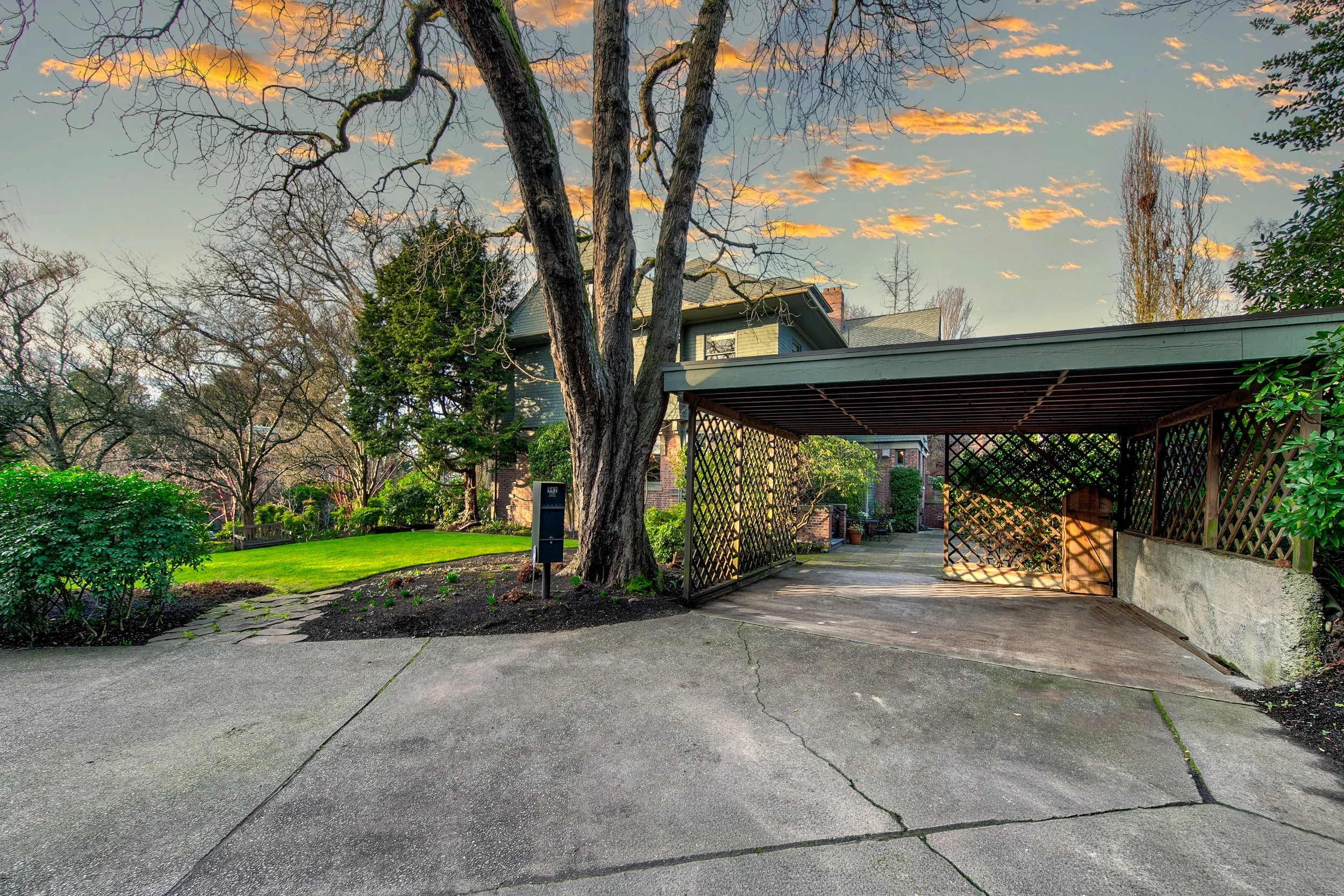 A driveway leading to a house with a carport on the right, surrounded by a well-maintained lawn and trees, at sunset with a partly cloudy sky.