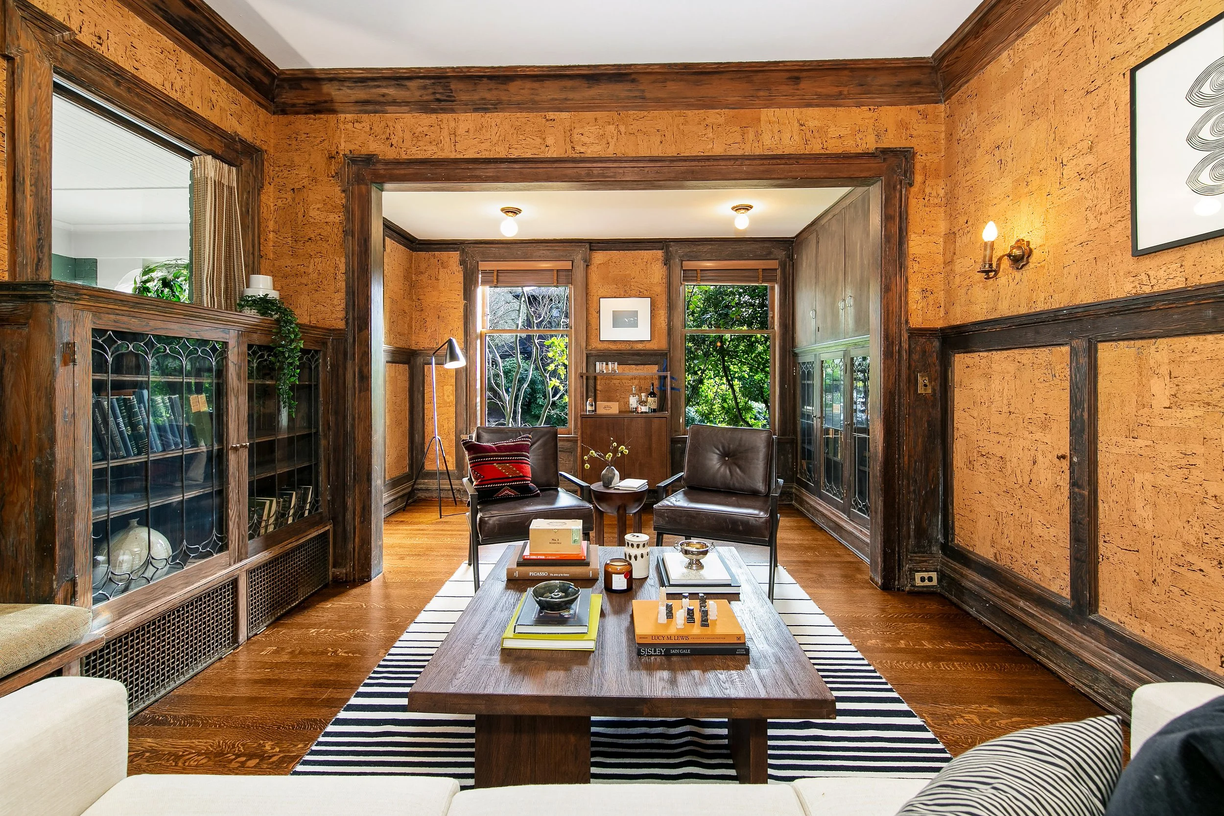 Living room with wood floors, vintage wooden walls, large windows with greenery outside, and mid-century modern furniture, including a coffee table, armchairs, and books.