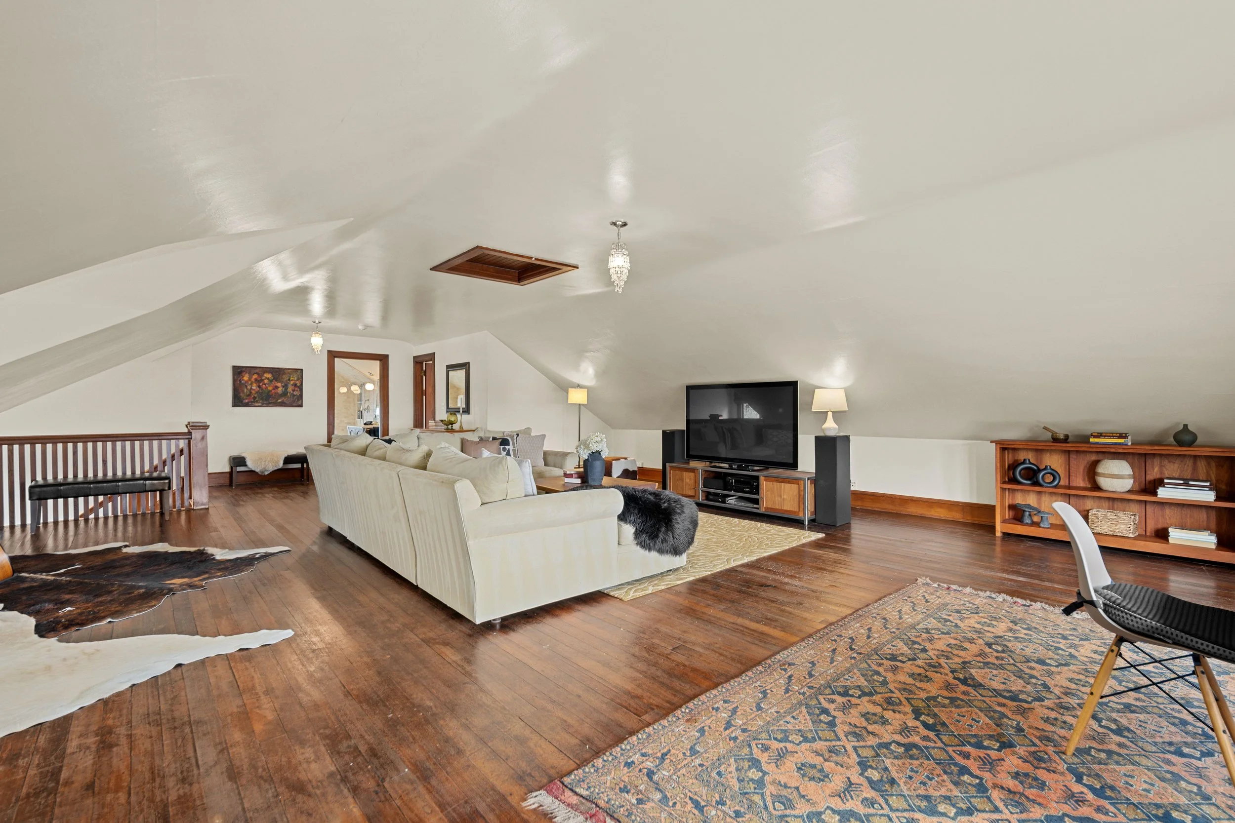 Living room with sloped ceiling, hardwood floors, beige sofa, black furry ottoman, large flat-screen TV on a wooden console, wooden bookshelf, area rug, and various lamps.