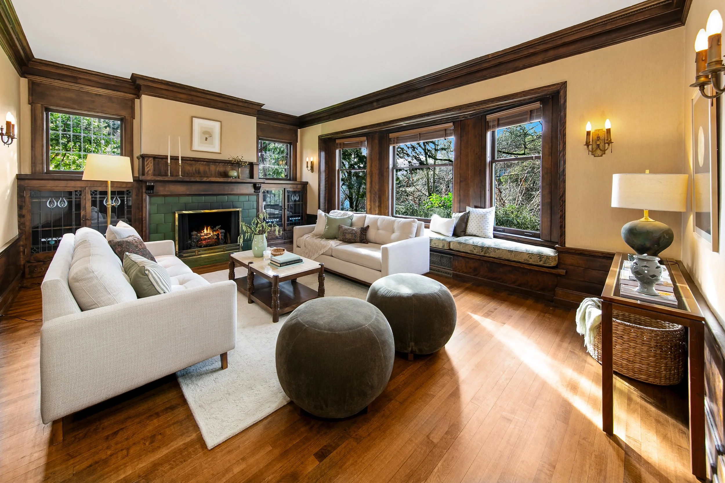 Living room with beige sofas, wood-paneled walls, large windows, bookshelf, fireplace, and decorative cushions.