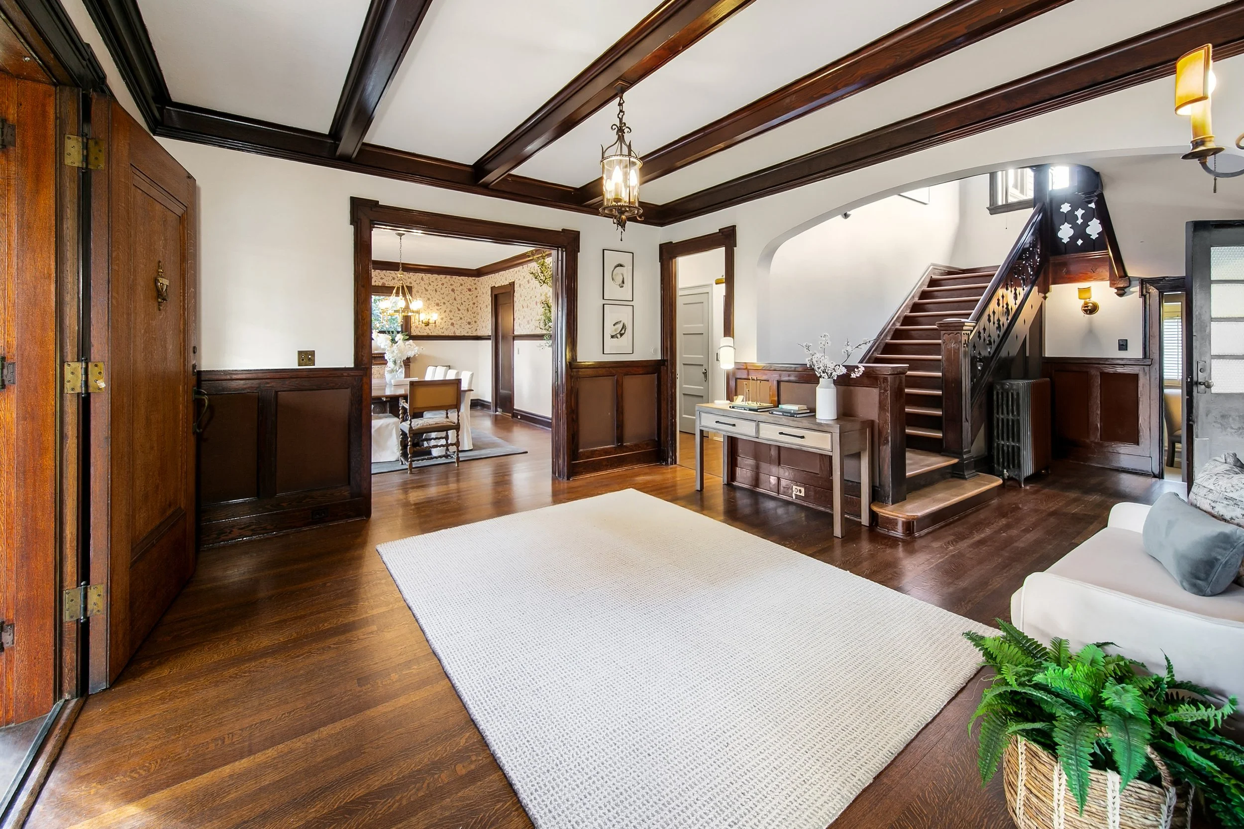 Living room with wood flooring, white walls with dark wood trim, staircase, white couch with gray pillows, a white side table with flowers, and a large beige rug.