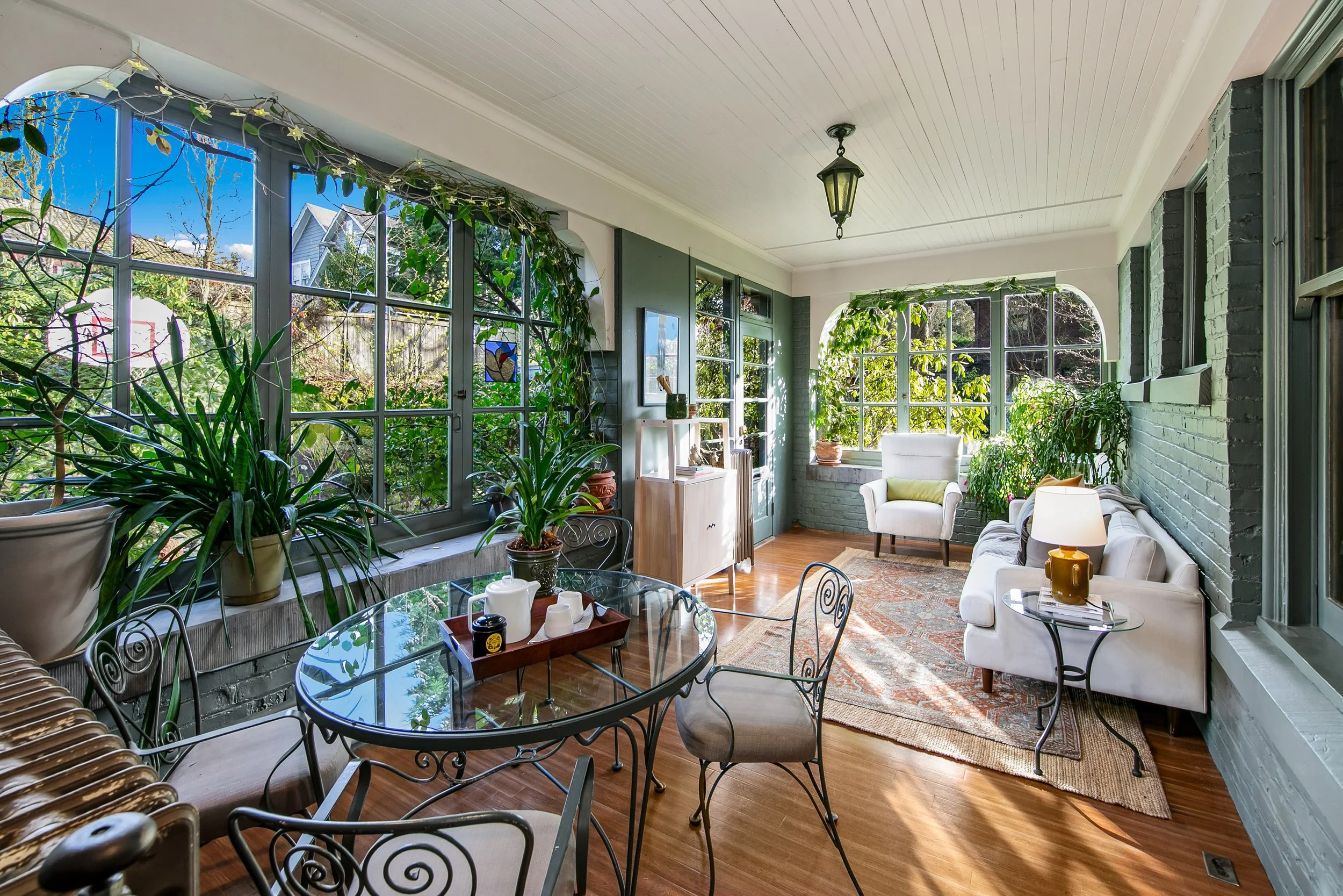 Cozy sunroom with large windows, green plants, white sofa, armchair, oval glass table, hardwood floor, and decorative rug, bright sunlight streams in.