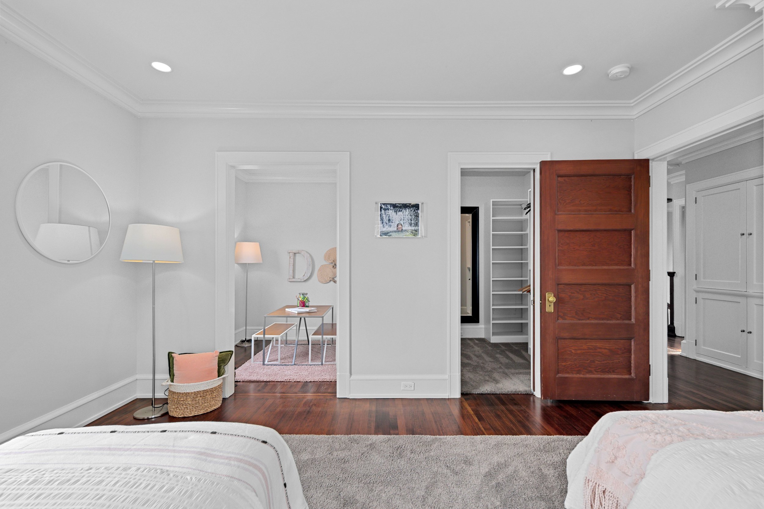 A bedroom with white walls and dark wood flooring. Two beds are partially visible at the bottom corners. To the left, a round mirror and a lamp stand beside a small basket. In the background, a small area with a table, two benches, and decorative wal