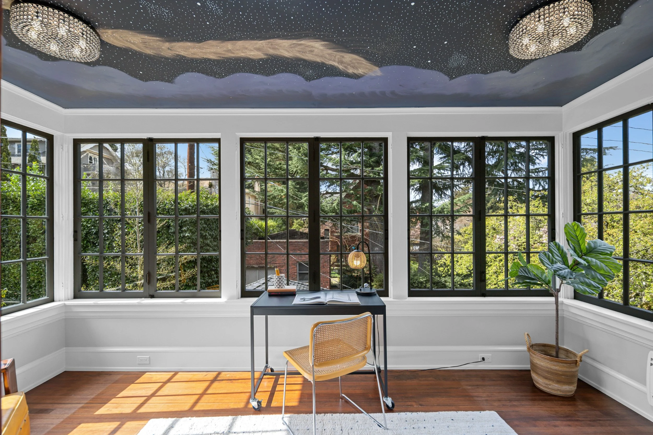 Bright sunlit room with large bay windows showing green trees outside. Room has a black desk with an open book, a lamp, and a pencil holder. There is a yellow chair in front of the desk, and a large potted plant with broad green leaves to the right. 
