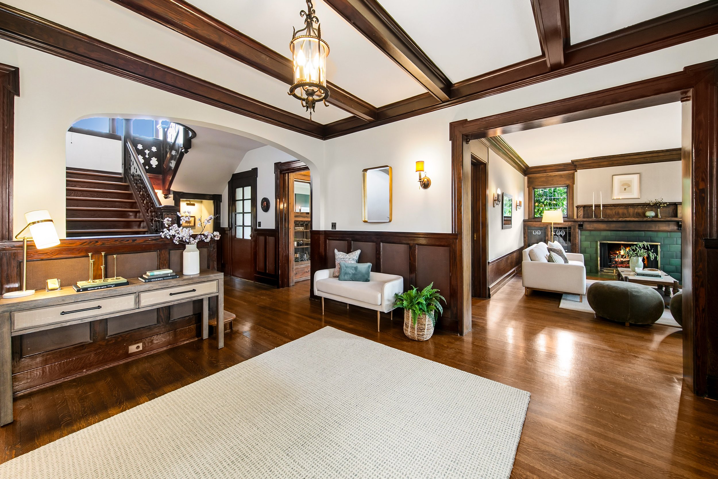 A spacious living room with wooden floors, white walls, and dark wood trim. There's a staircase with a dark wood banister on the left, and a doorway leading to another room with a fireplace and green tile. The room is decorated with a white bench, a 