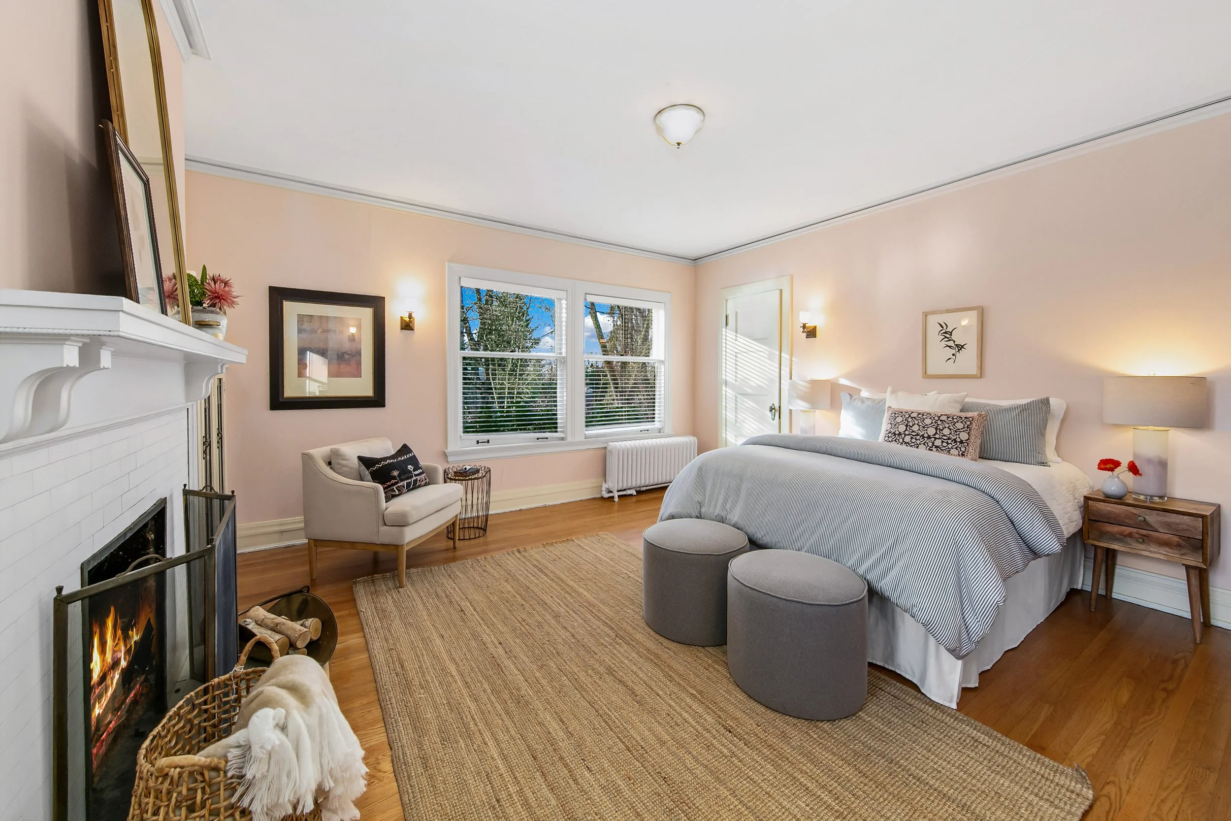 A bedroom with a large bed, bedside tables with lamps, a fireplace, a chair near the window, and two gray ottomans at the foot of the bed.