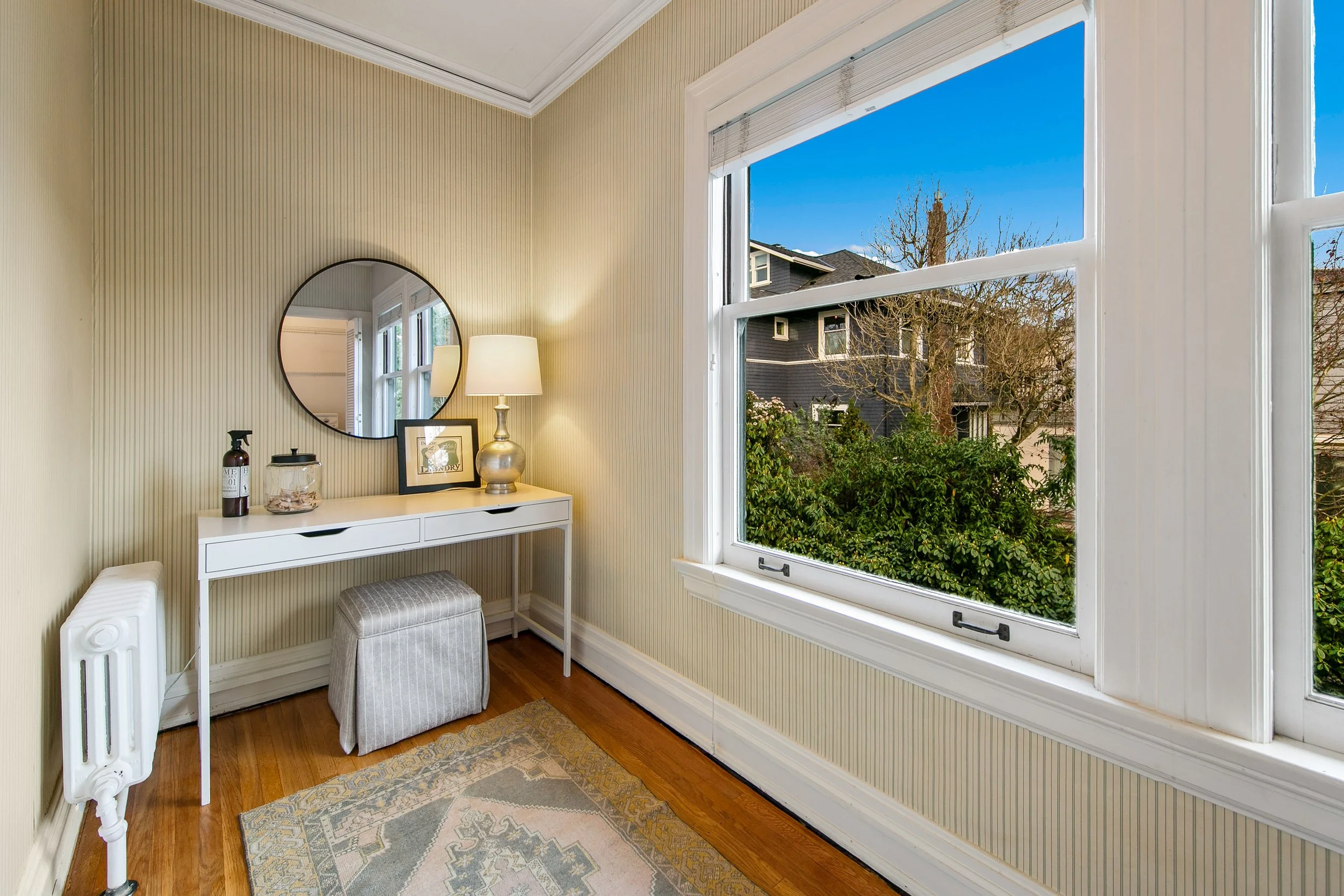 A cozy corner of a room with a white desk, a mirror, a table lamp, framed photos, and decorative items. There is a window showing a view of trees and neighboring houses outside.