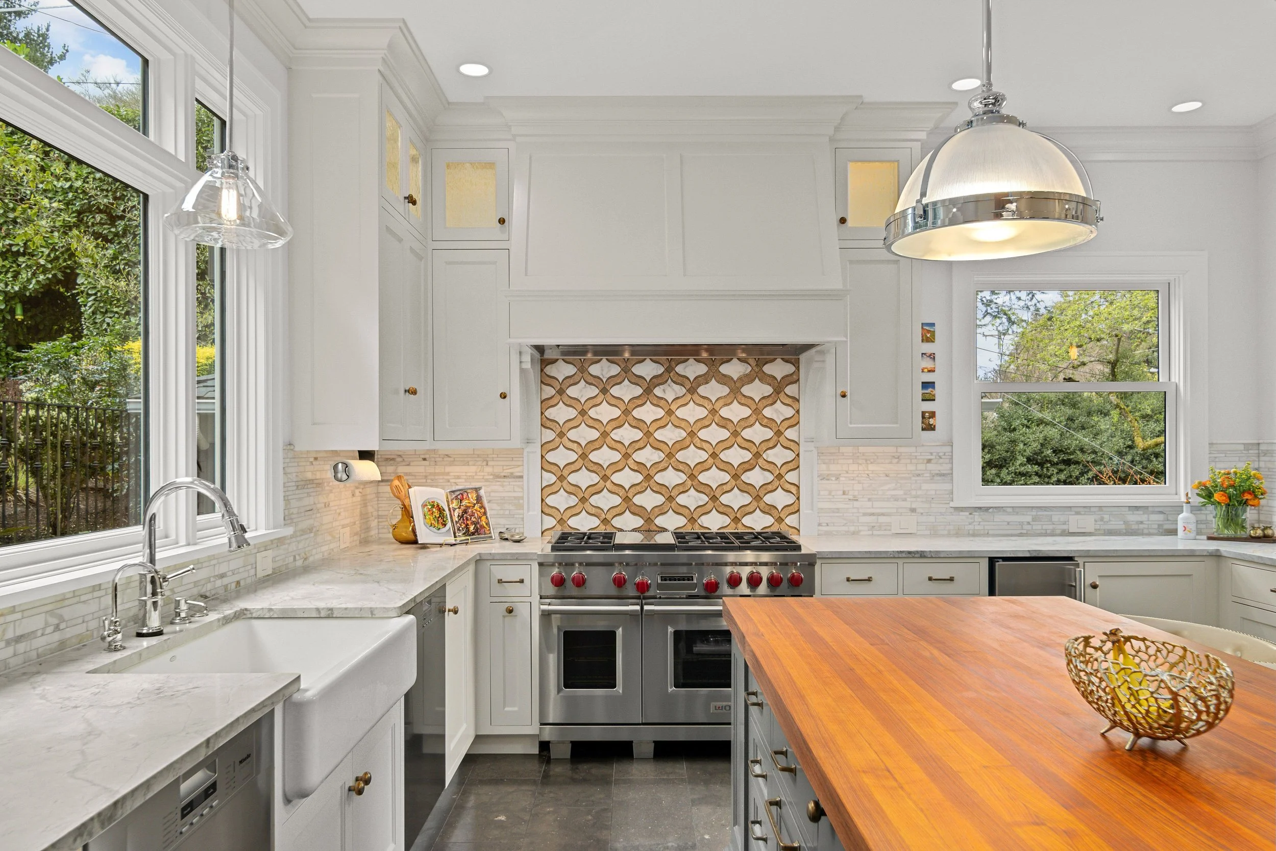 Modern kitchen with white cabinets, marble countertops, a double oven, large windows showing greenery outside, a wooden kitchen island, and decorative backsplash above the stove.