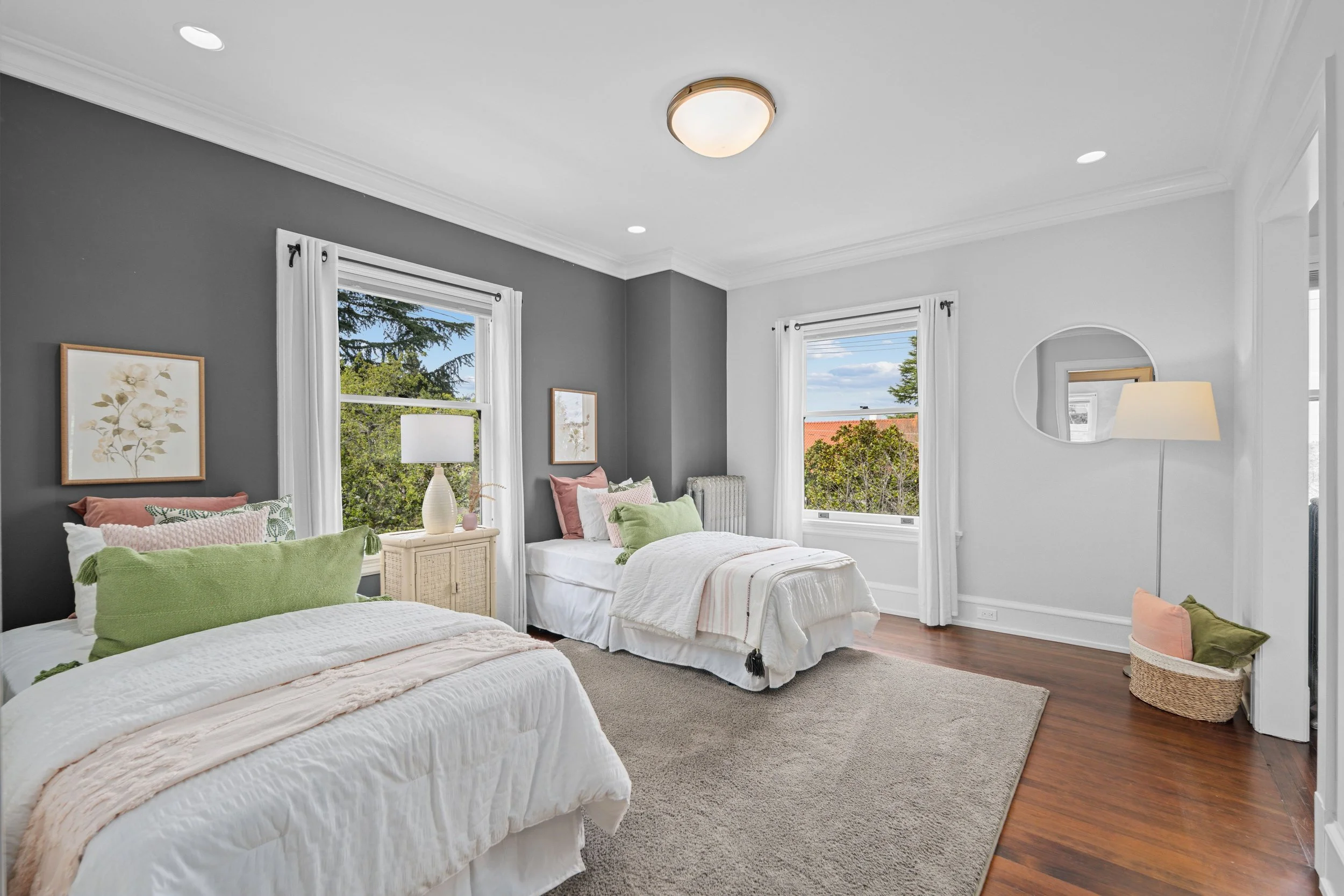 A bright bedroom with two white beds, green and pink pillows, a beige nightstand with a lamp, large windows with white curtains, a round wall mirror, a floor lamp, a woven basket, and artwork on a gray accent wall.