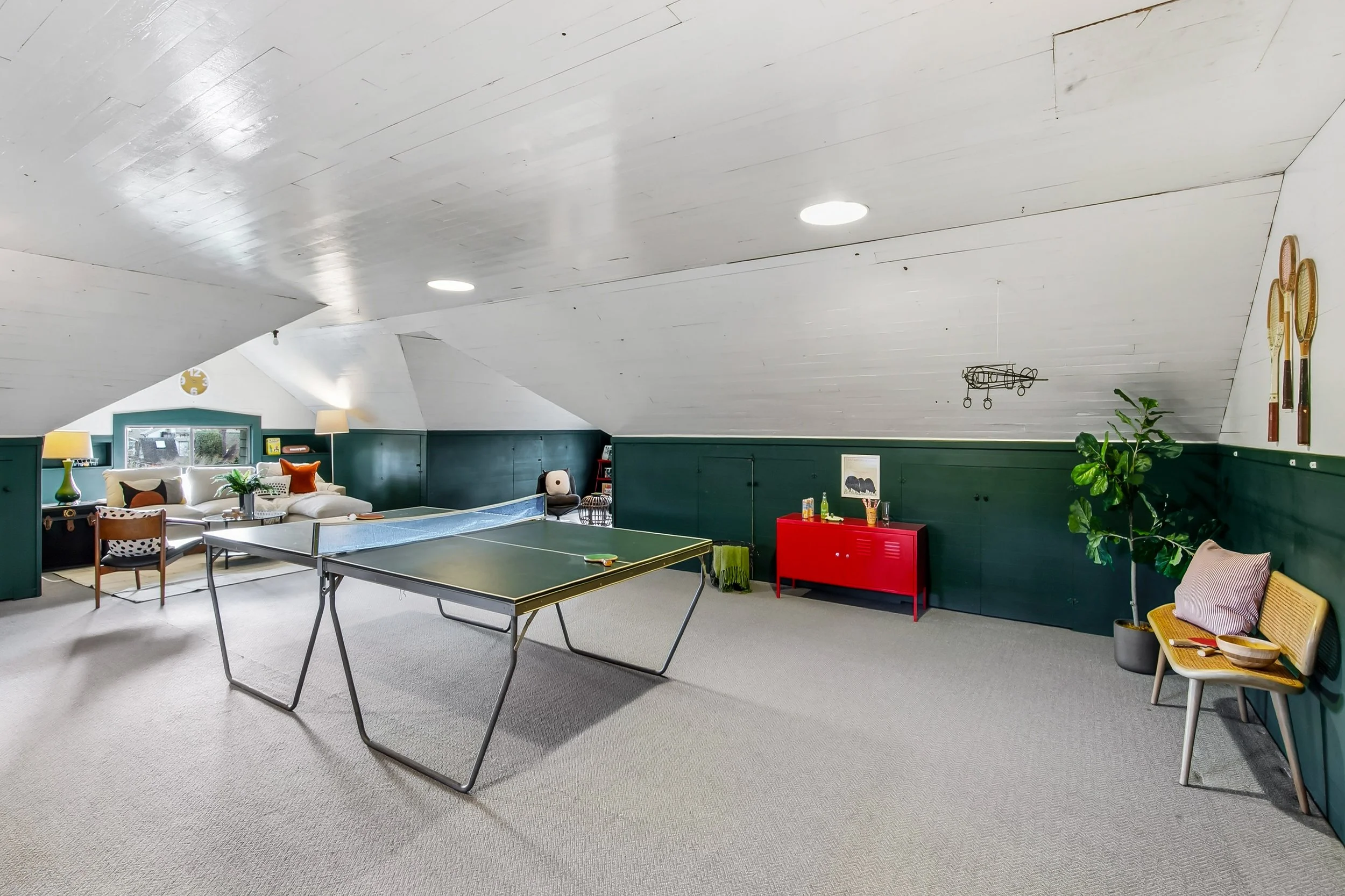 An attic game room with a ping pong table in the center, a sofa with cushions, a side table with a lamp, a red cabinet, and various decorative items and tennis rackets on the wall.