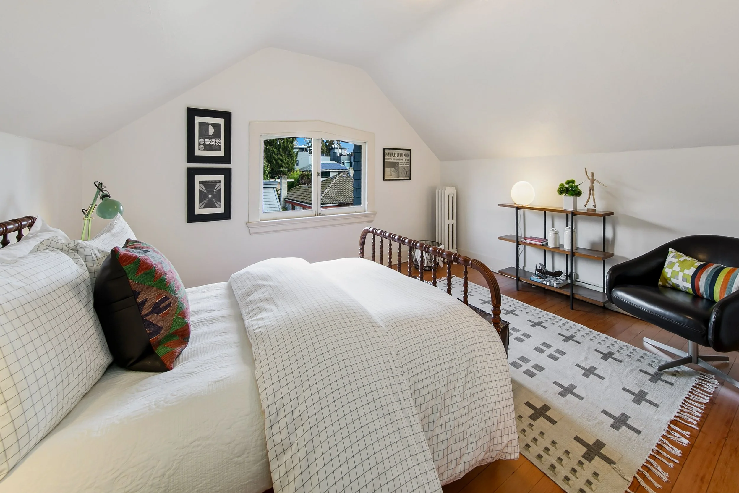 A cozy bedroom with a white bed, a black armchair, a wooden shelf with a lamp and decor, and a window showing a view of rooftops and trees.