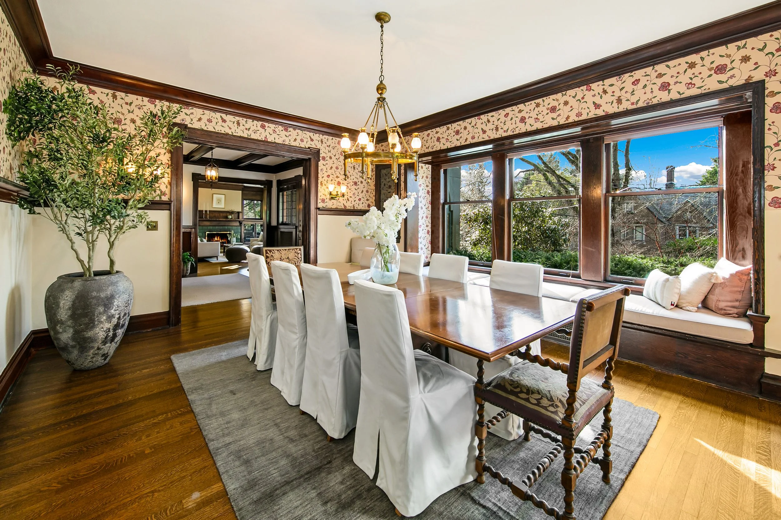 Bright dining room with large windows, a wooden table surrounded by eight white slipcovered chairs, a large potted plant, and a floral wallpapered wall.