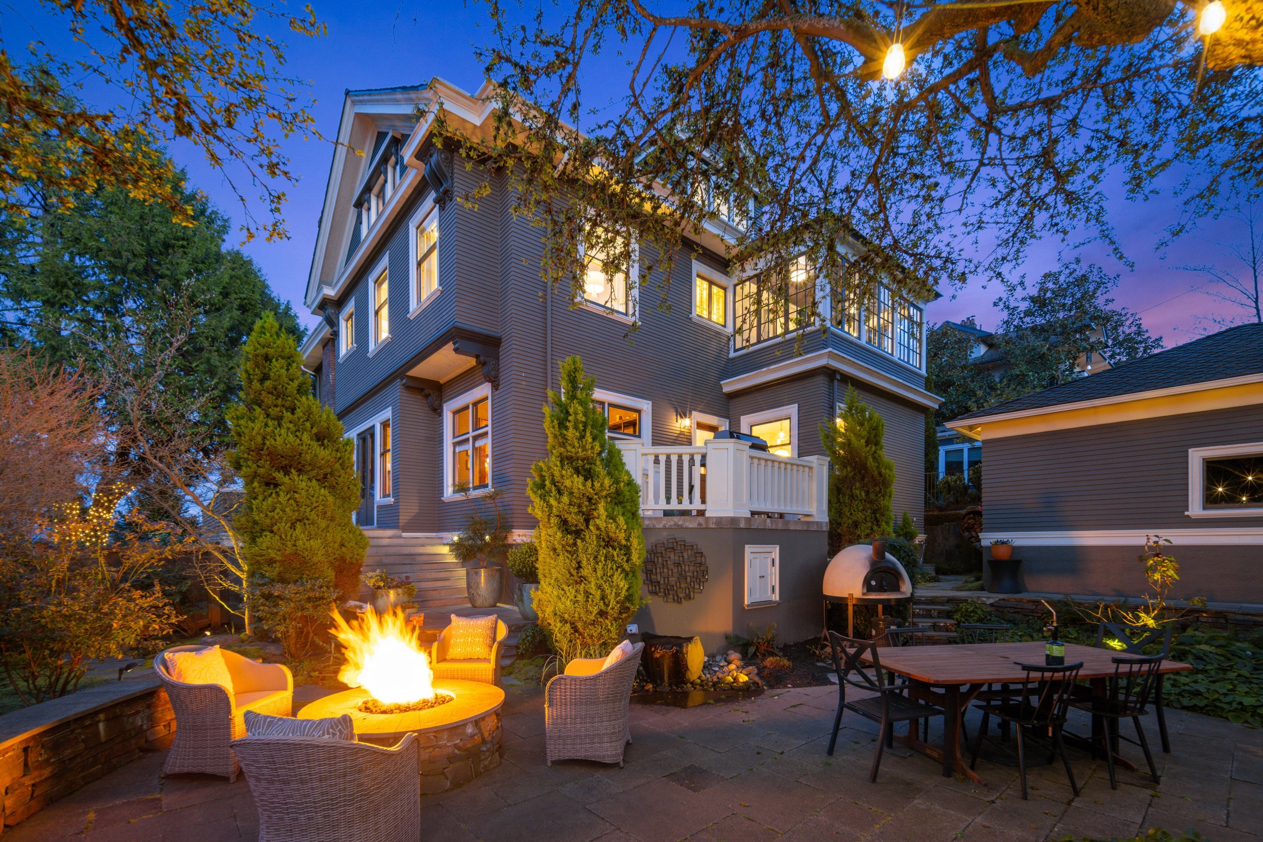 A multi-story gray house with lit windows, outdoor patio with fire pit, seating, dining table, pizza oven, and surrounding trees during twilight.