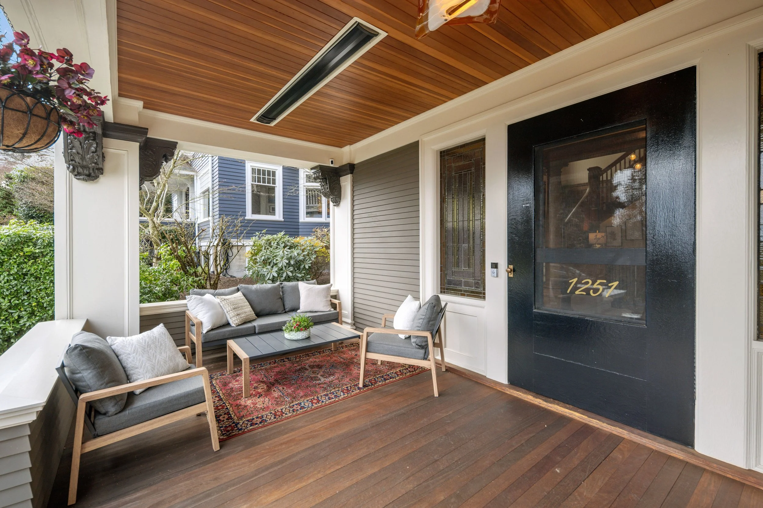 Porch with wooden floor and ceiling, black door with house number 1257, gray cushioned chairs, a gray sofa with pillows, a rectangular black coffee table, a red patterned rug, and greenery outside.