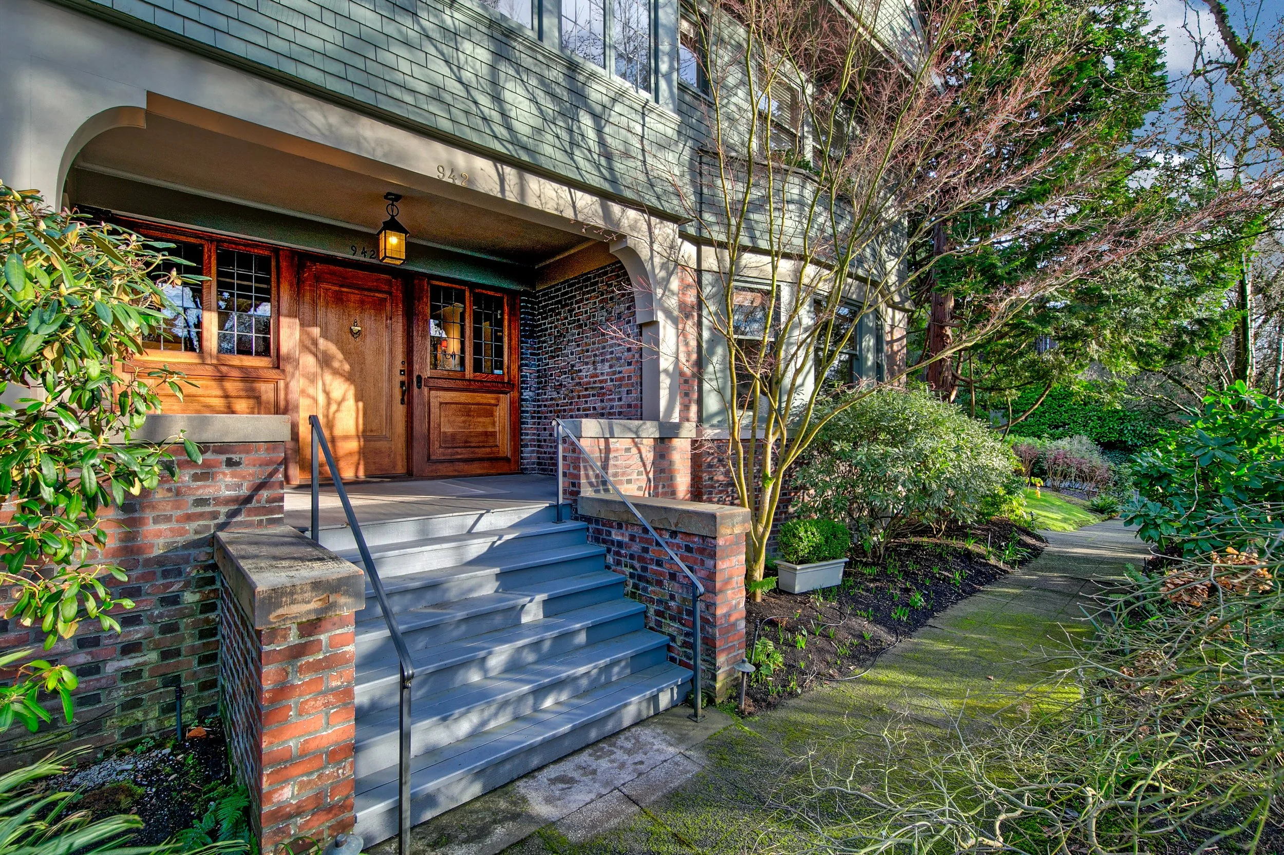 Front porch of a house with brick stairs, wooden door, and a lantern light, surrounded by lush greenery and a paved walkway.