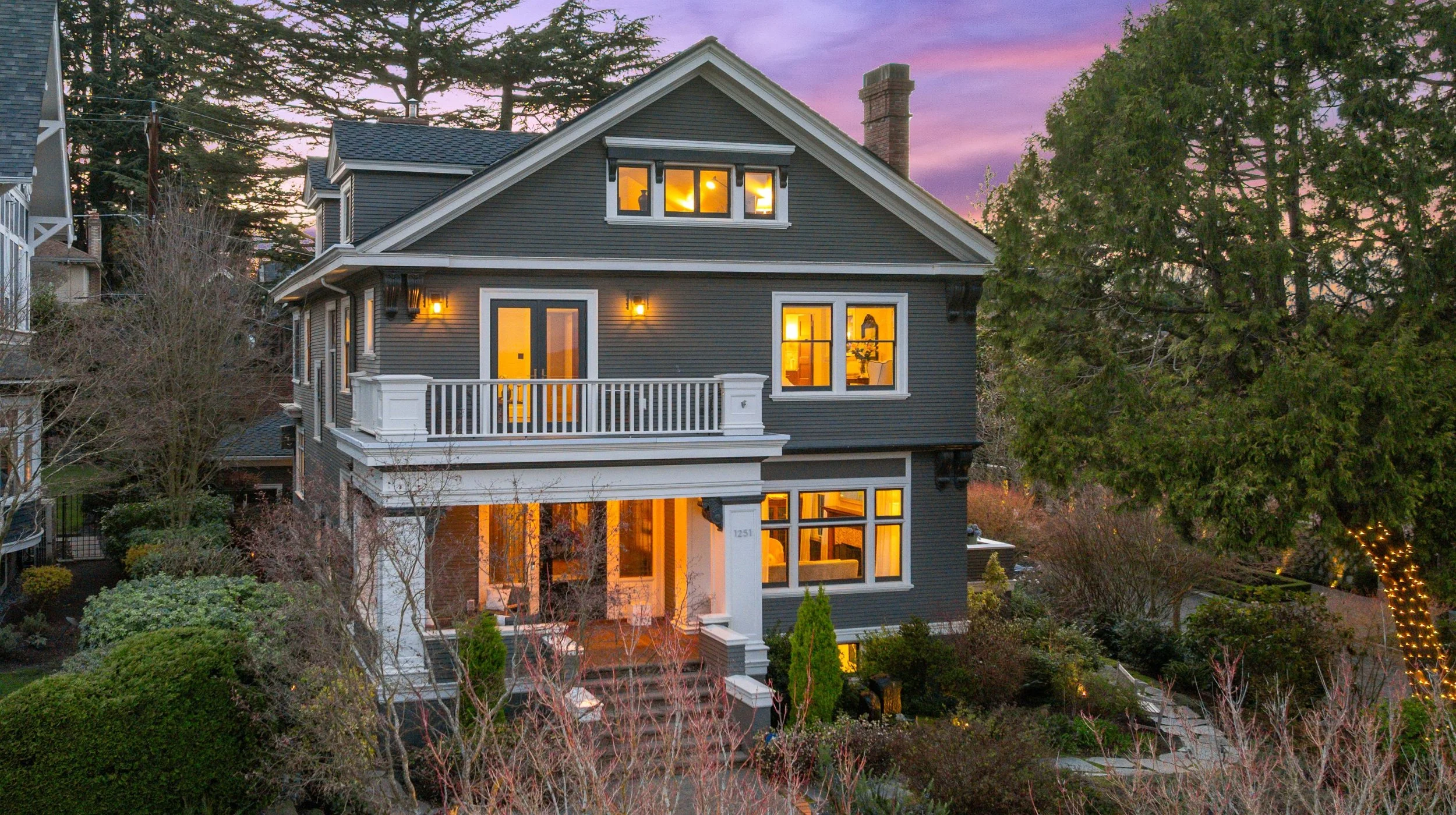 A multi-story house with dark gray siding and white trim lit from within, with a porch and balcony, surrounded by trees and garden at sunset, with a purple sky in the background.