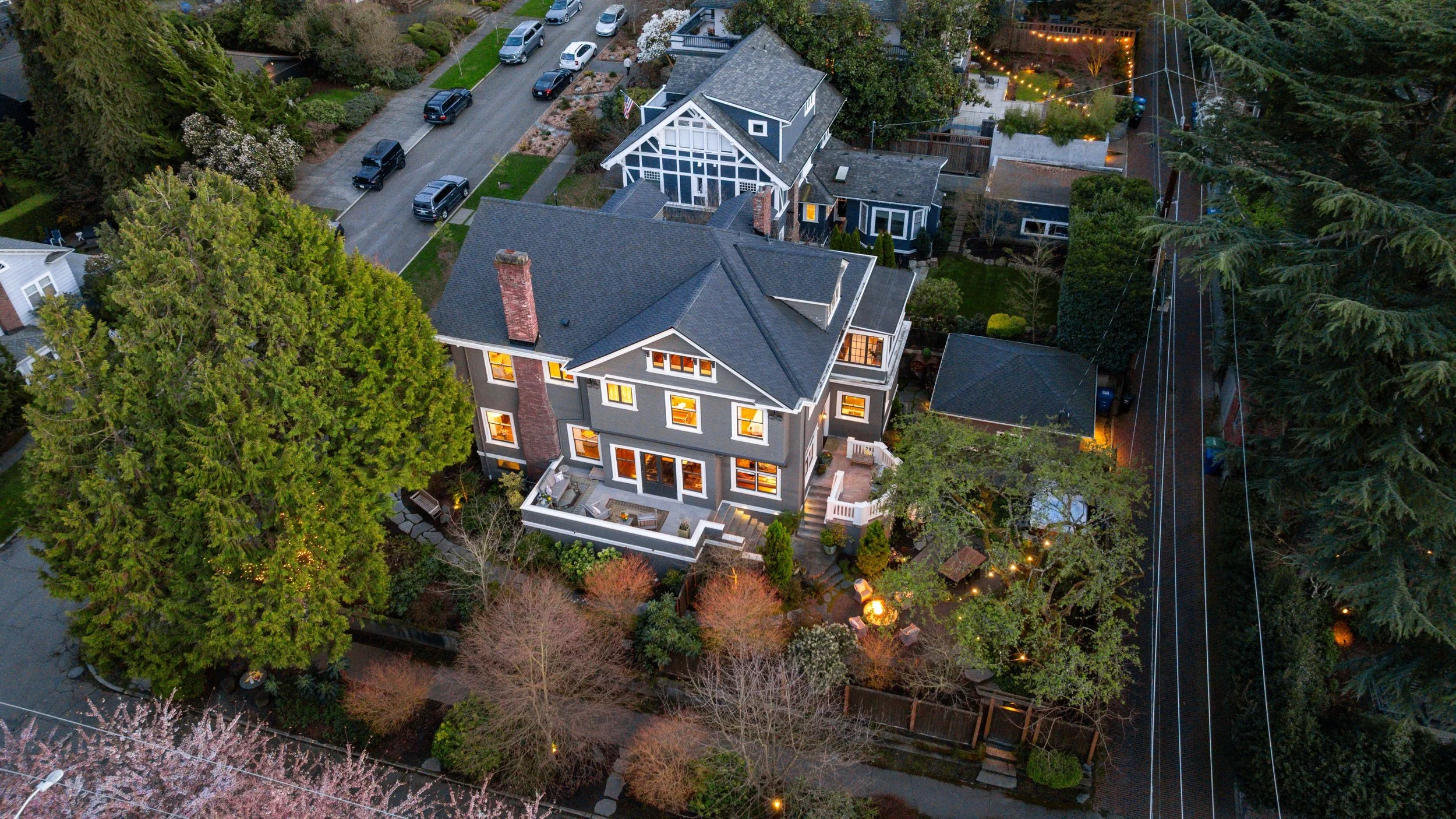 Aerial view of a large gray house with white trim, surrounded by trees and a garden, with a street and parked cars nearby at dusk.