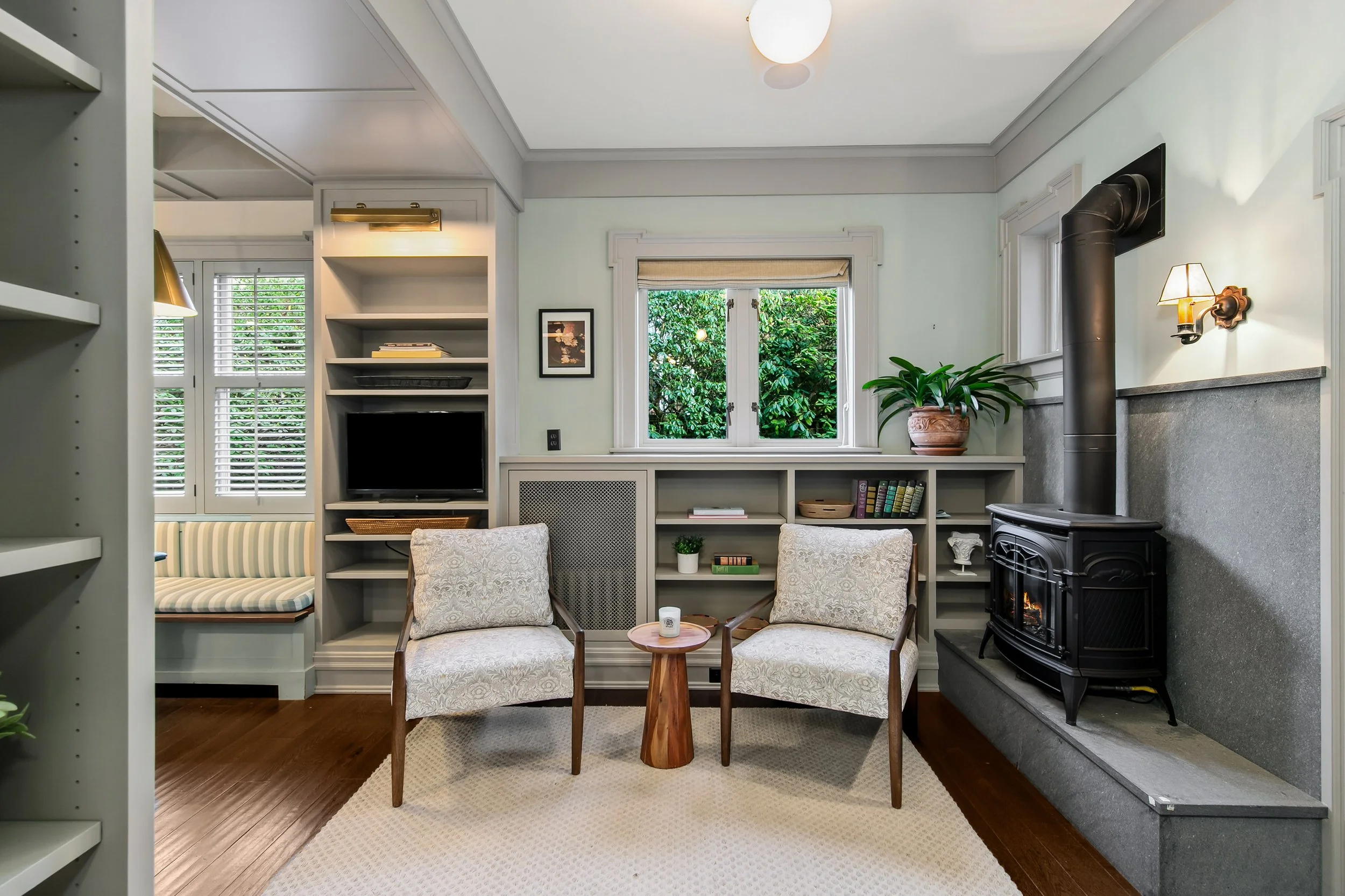 Cozy living room with two armchairs, a small wooden side table, a fireplace with a black wood stove, and windows with greenery outside.