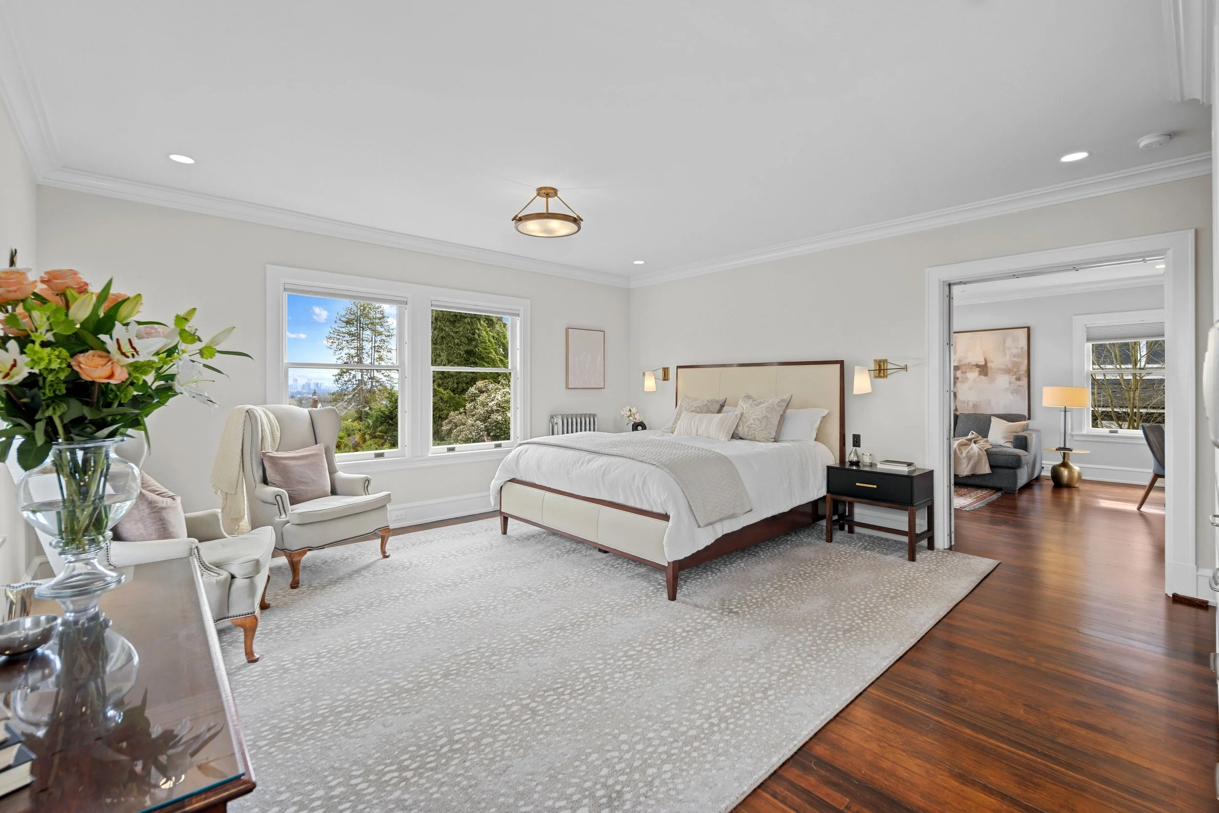 Bright bedroom with white walls, large windows showing trees and city view, white bedding, decorative pillows, bedside lamps, and a sitting area visible through doorway.