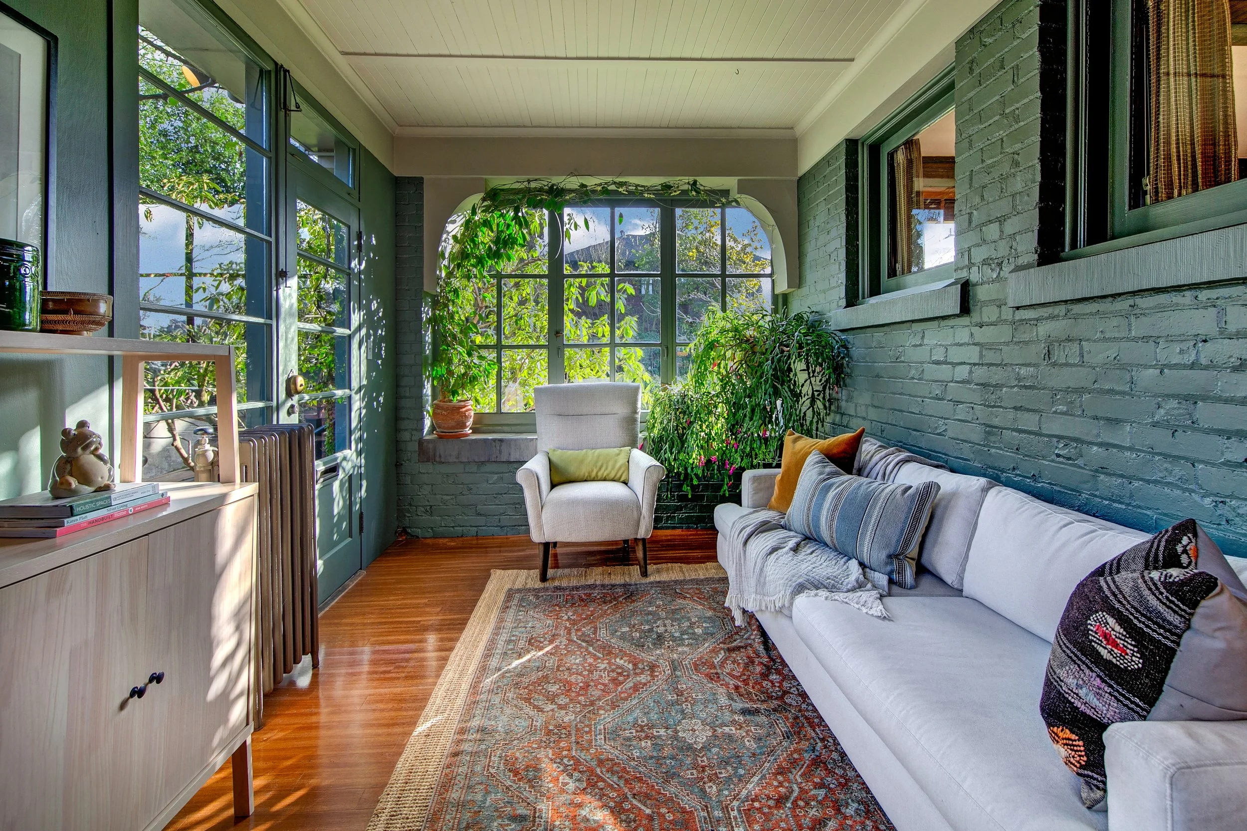 Bright living room with large windows, green brick walls, a white armchair with a yellow pillow, a white sofa with multiple colorful pillows, a patterned rug, indoor plants, and wooden flooring.