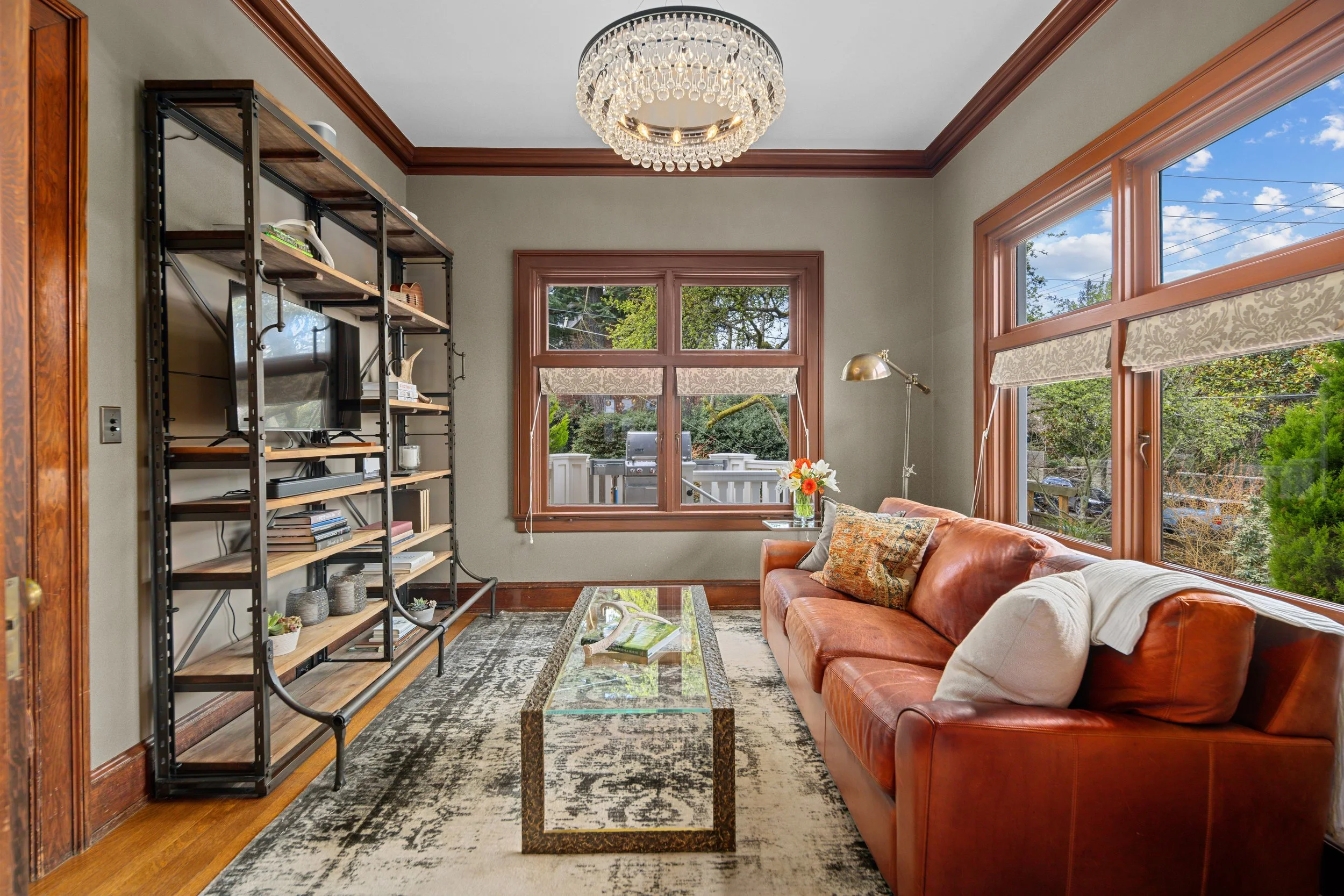 Living room with large windows, a leather couch, a glass coffee table, a metal shelving unit, a window with blinds, and a chandelier.