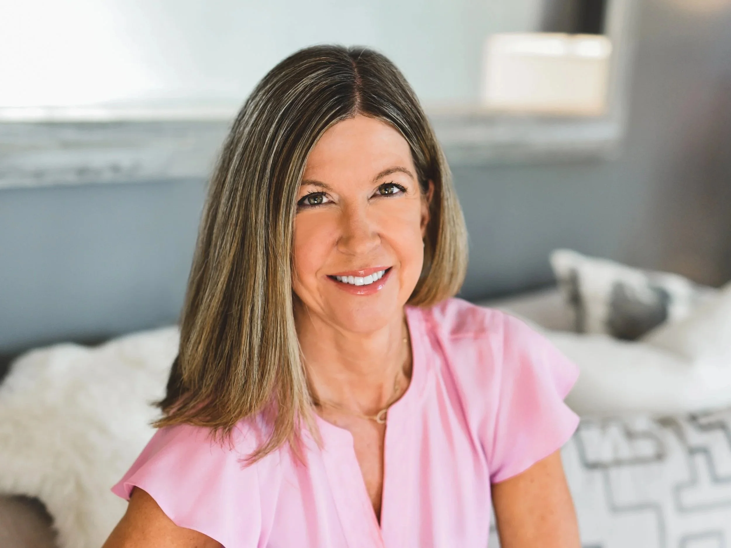 A smiling woman with shoulder-length light brown hair, wearing a pink top, sitting on a bed in a bright room.