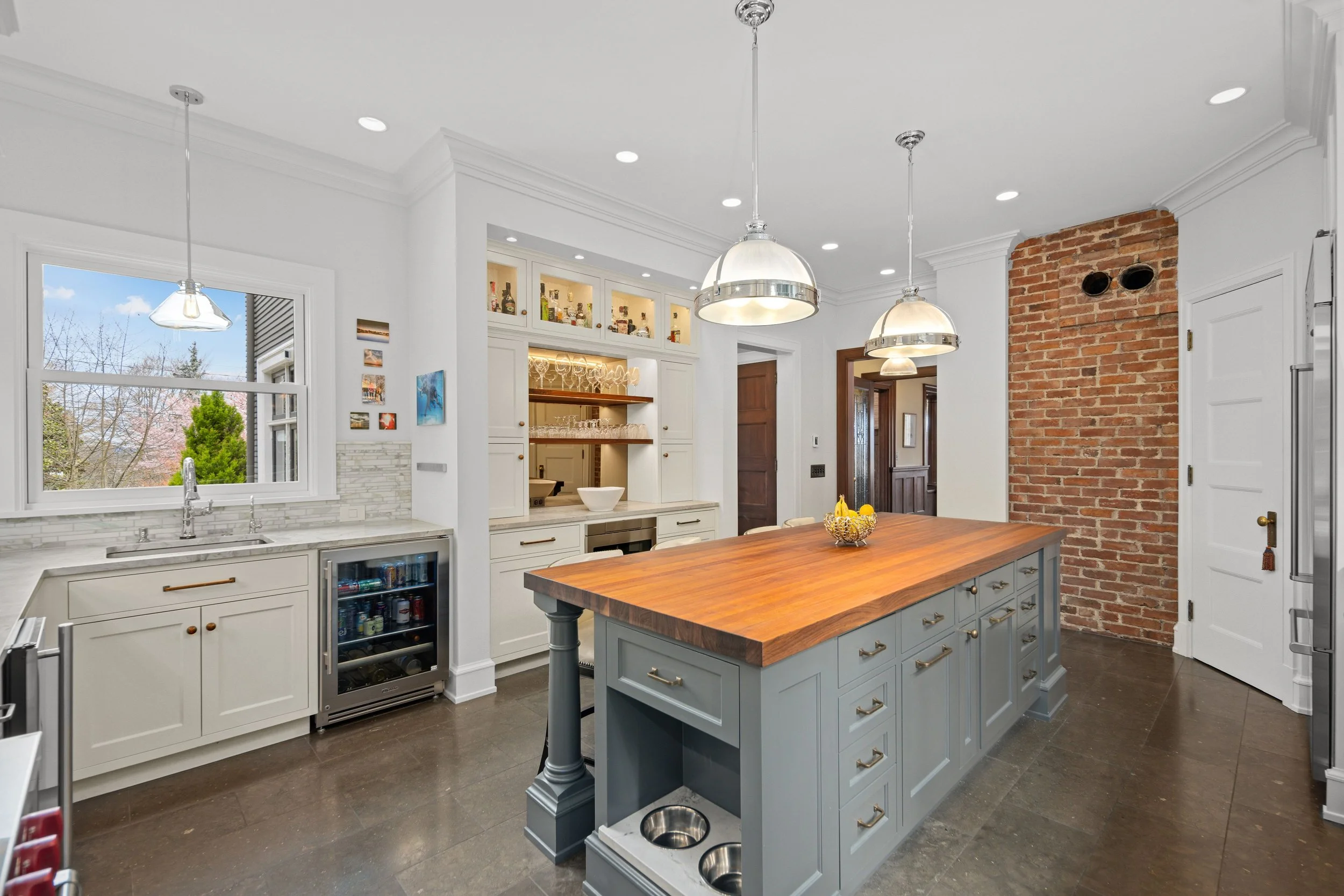 Modern kitchen with white cabinets, a brick accent wall, large window, wooden island with gray base, pendant lighting, and dark floor.