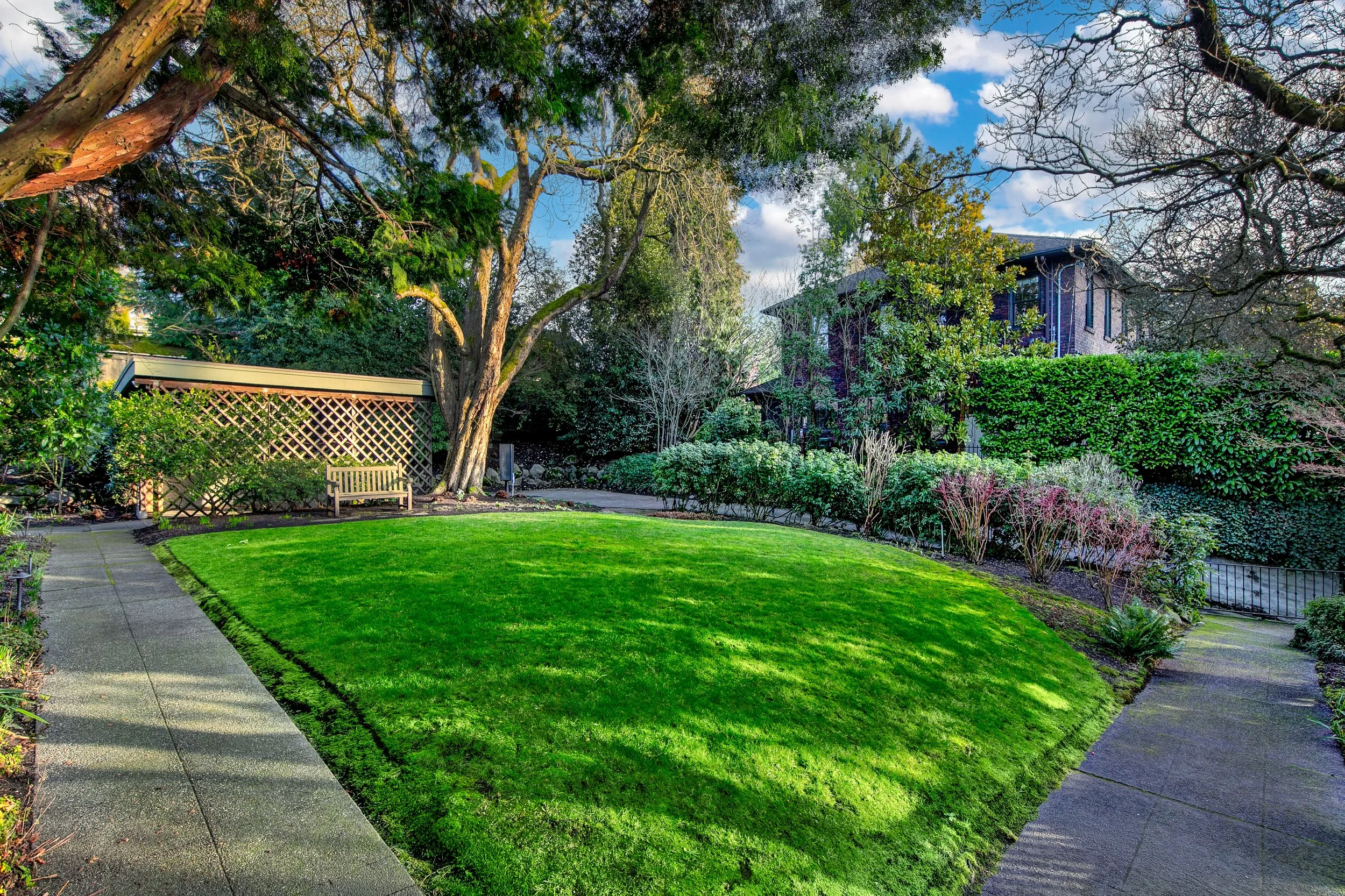 A lush green backyard with a neatly trimmed grass lawn, surrounded by various trees and shrubs. A paved walkway runs along the edge, leading to a wooden bench near a small garden shed. There is a multi-story house in the background partially obscured