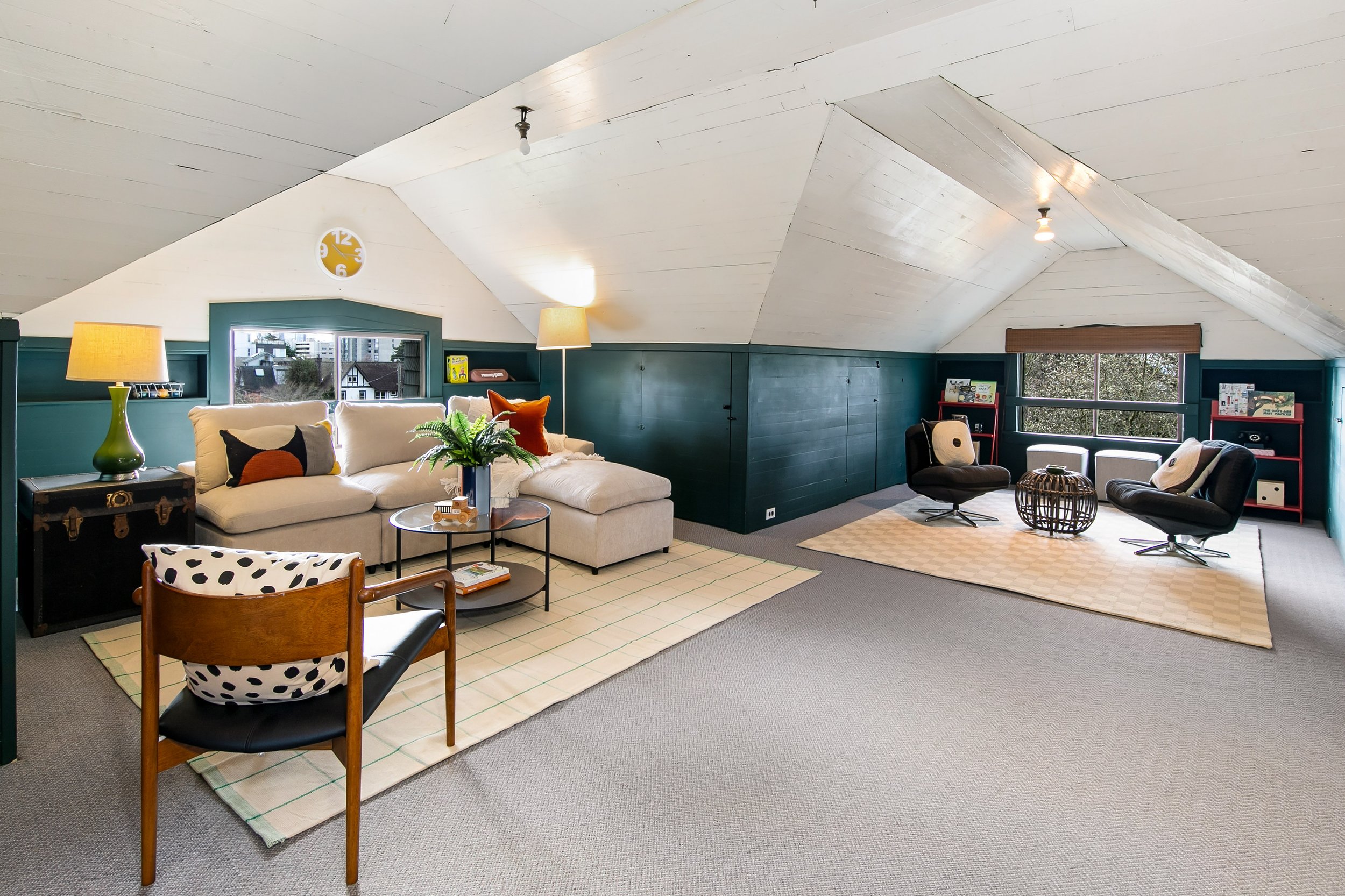 Cozy attic living room with white ceiling, teal walls, beige sofa, black chairs, and decorative pillows, illuminated by lamps and natural light from windows.
