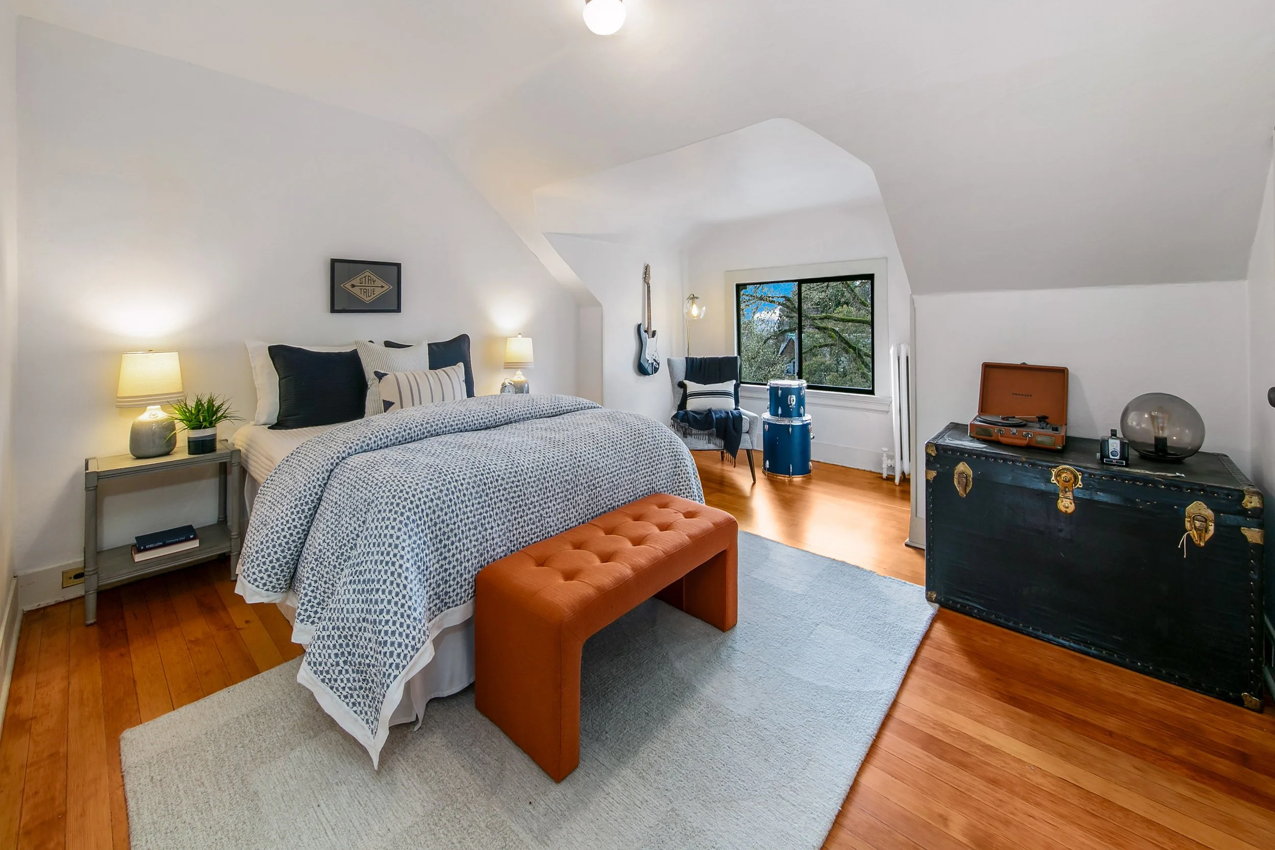 Bedroom with white walls and sloped ceiling, wooden floor, and a large bed with black and white pillows. Next to the bed are two lamps and a small nightstand with a plant. At the foot of the bed is an orange tufted bench. In the corner near a window,