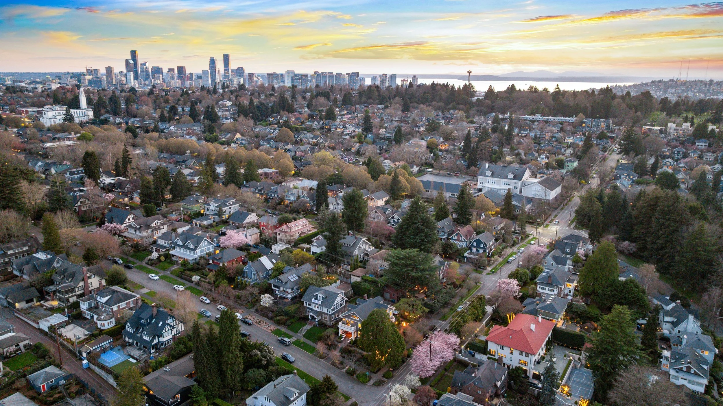 Aerial view of a city neighborhood during sunset, showing a mix of houses, trees, and streets with a downtown skyline in the background and a body of water beyond.