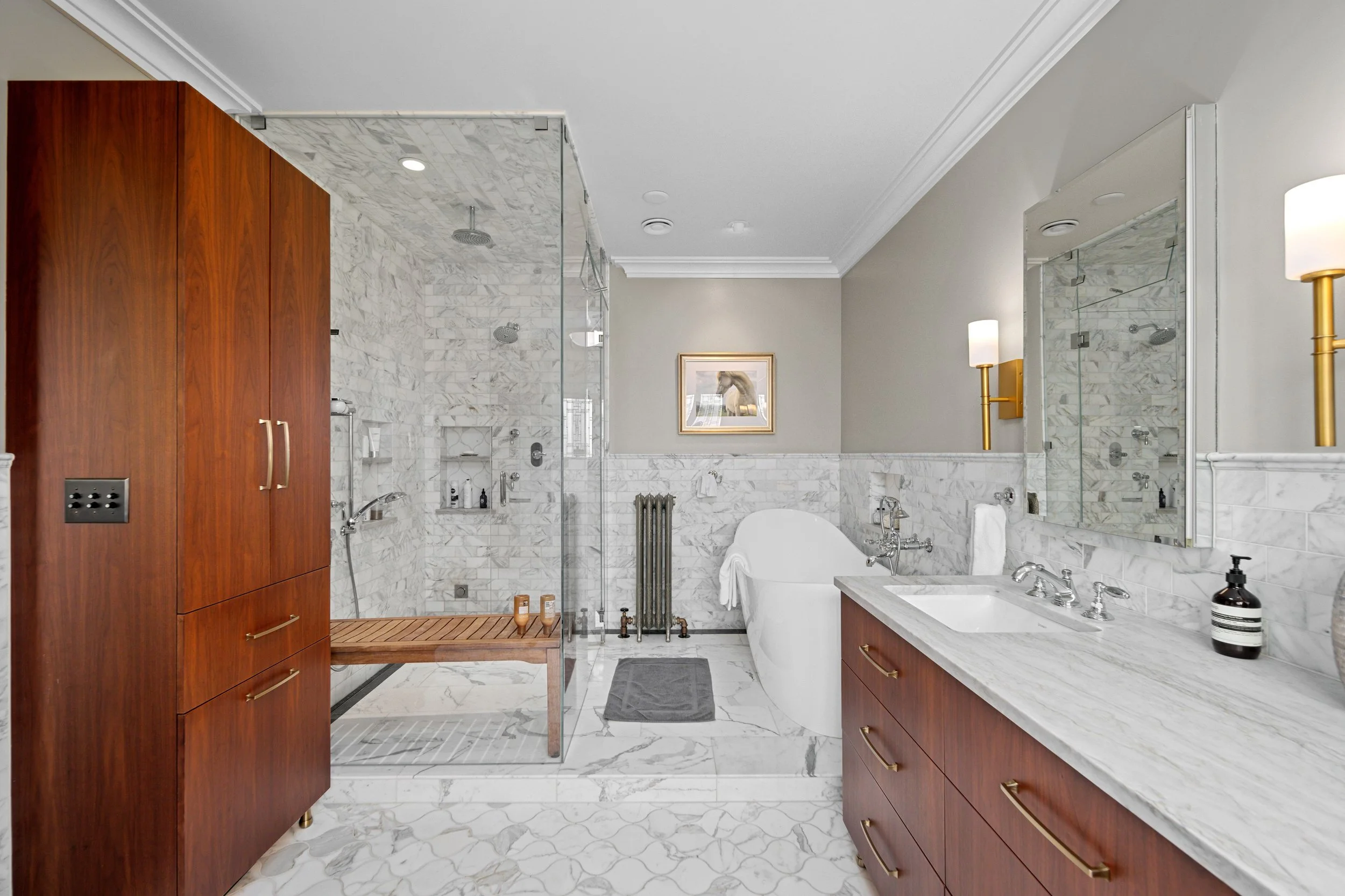 A luxurious bathroom with a marble floor and walls, a glass-enclosed shower area, a wooden cabinet, a white freestanding bathtub, mirror, and brass wall-mounted lights.