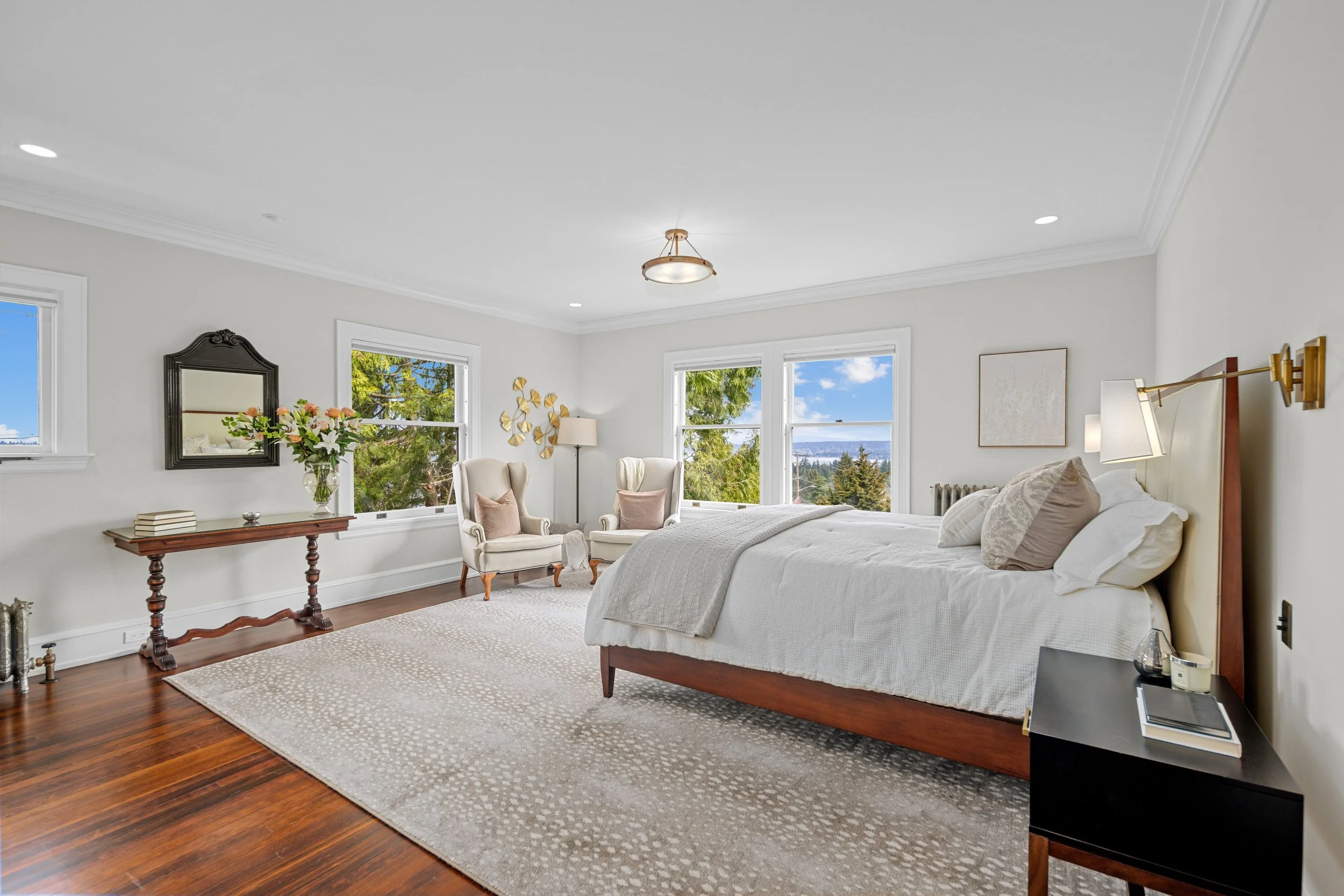 Bright bedroom with white walls, wooden floor, large bed with white bedding, two white armchairs with pink cushions near windows, bedside table with books and glass, mirror on wall, and view of trees and blue sky outside