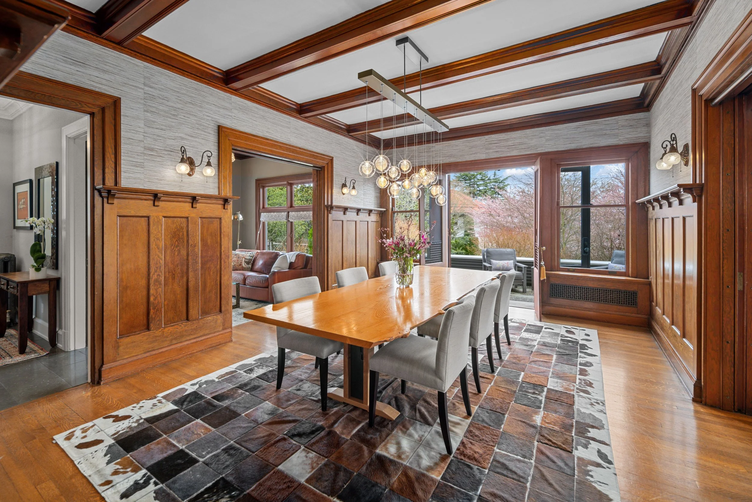 A dining room with a wooden table and gray upholstered chairs, large windows overlooking a backyard with trees, and a hanging modern light fixture with multiple globes.
