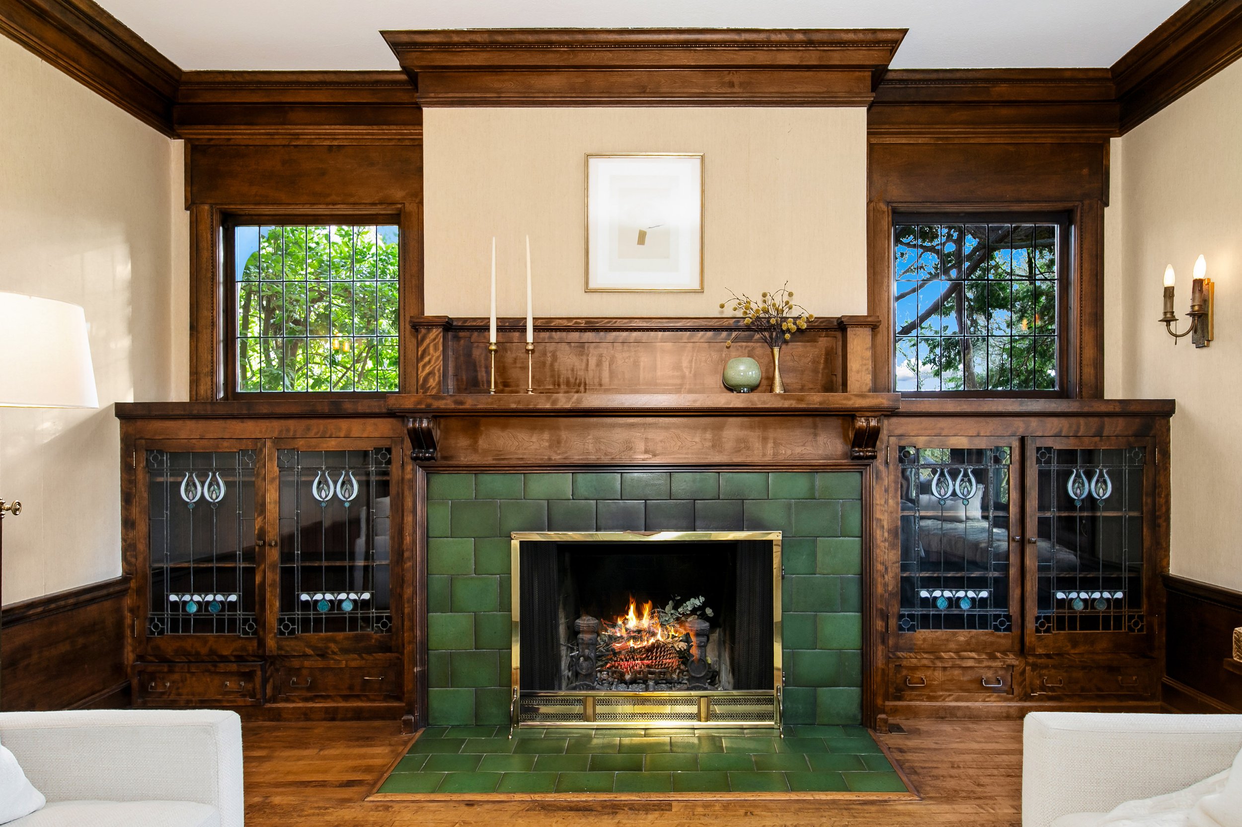 Living room with a green tiled fireplace, wooden mantel, two windows with grid panes, and built-in wooden cabinets on either side. Decorations include a framed picture above the mantel, white candles, a vase with branches, and a small vase with flowe
