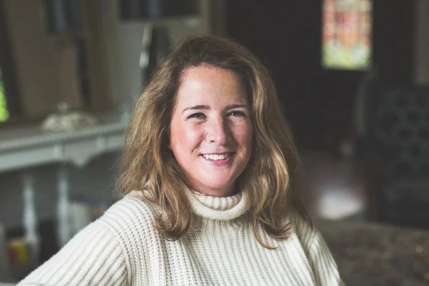 A woman with shoulder-length light brown hair smiling, wearing a beige knit sweater, indoors with a blurred background.