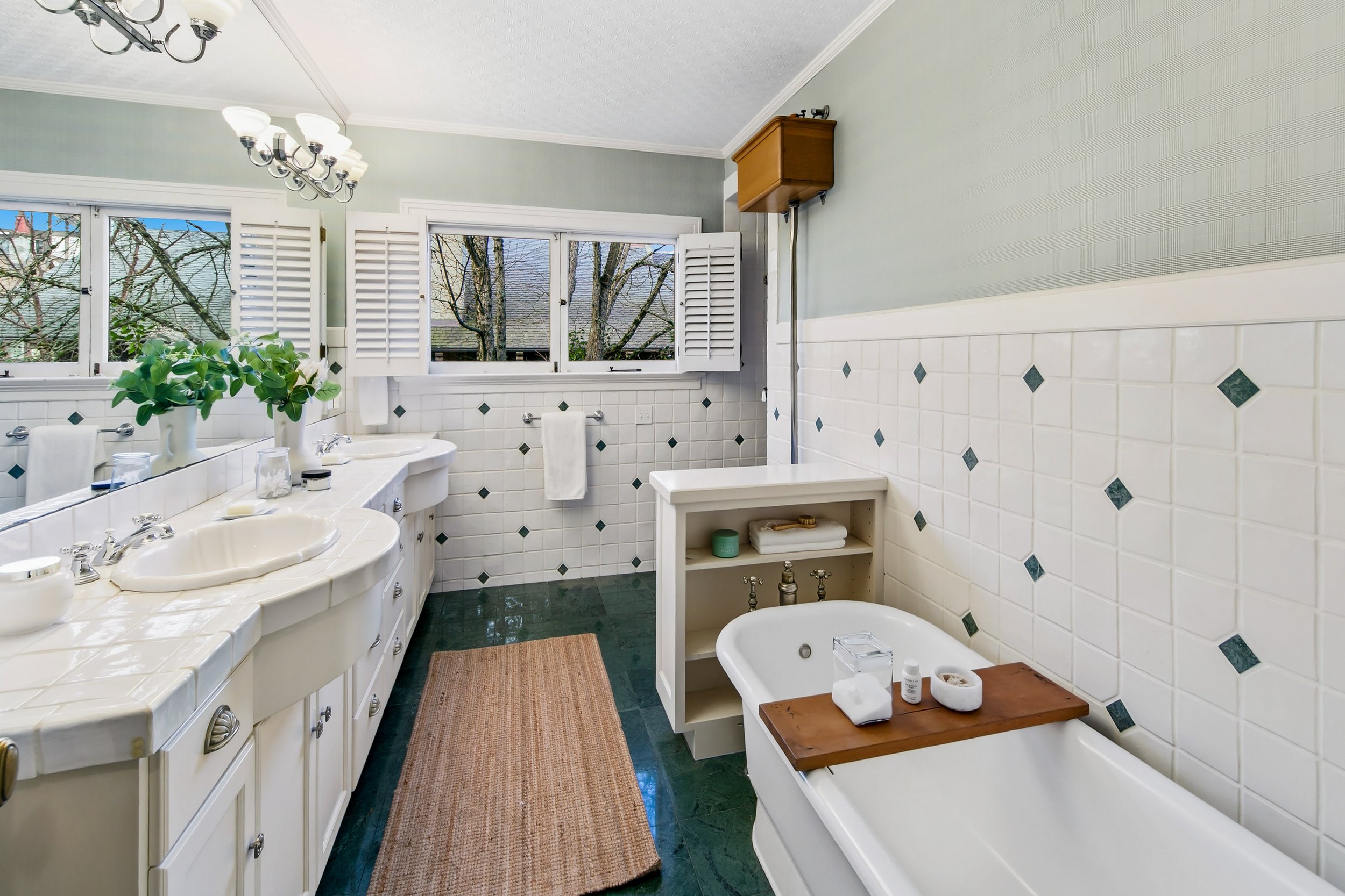 A bathroom with white tiled walls and green tiled floor, featuring a bathtub, double sink vanity with mirror, and open window with shutters showing trees outside.