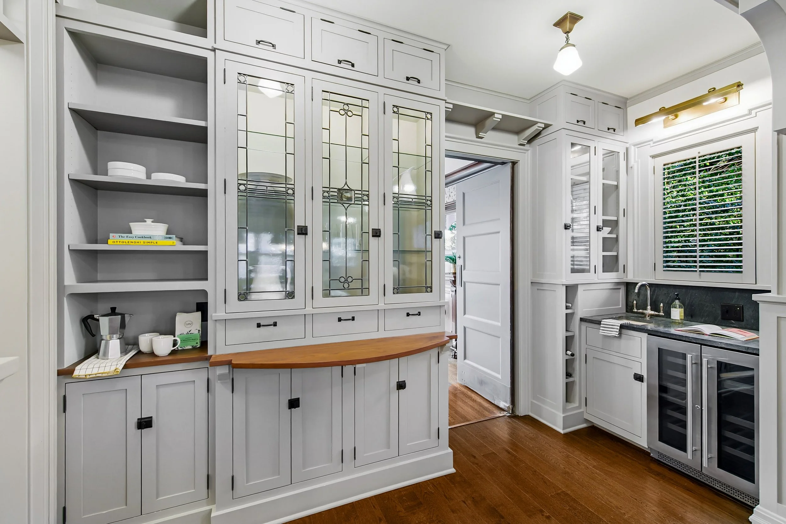 A kitchen with white cabinetry, a small wine cooler, a window with blinds, and open shelves with dishes and books.