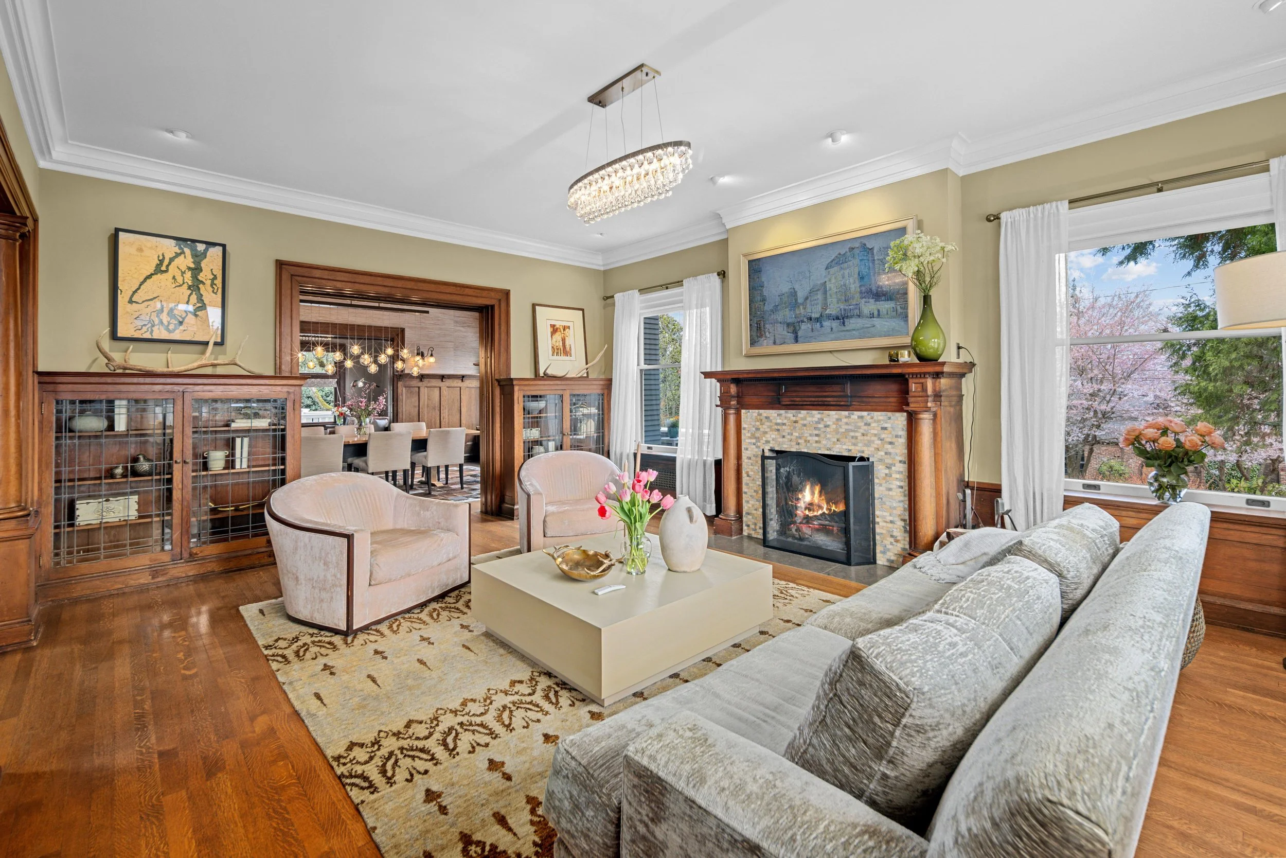 Living room with a fireplace, beige sofa, pink velvet armchairs, a coffee table with pink tulips, window with white curtains, and a chandelier.