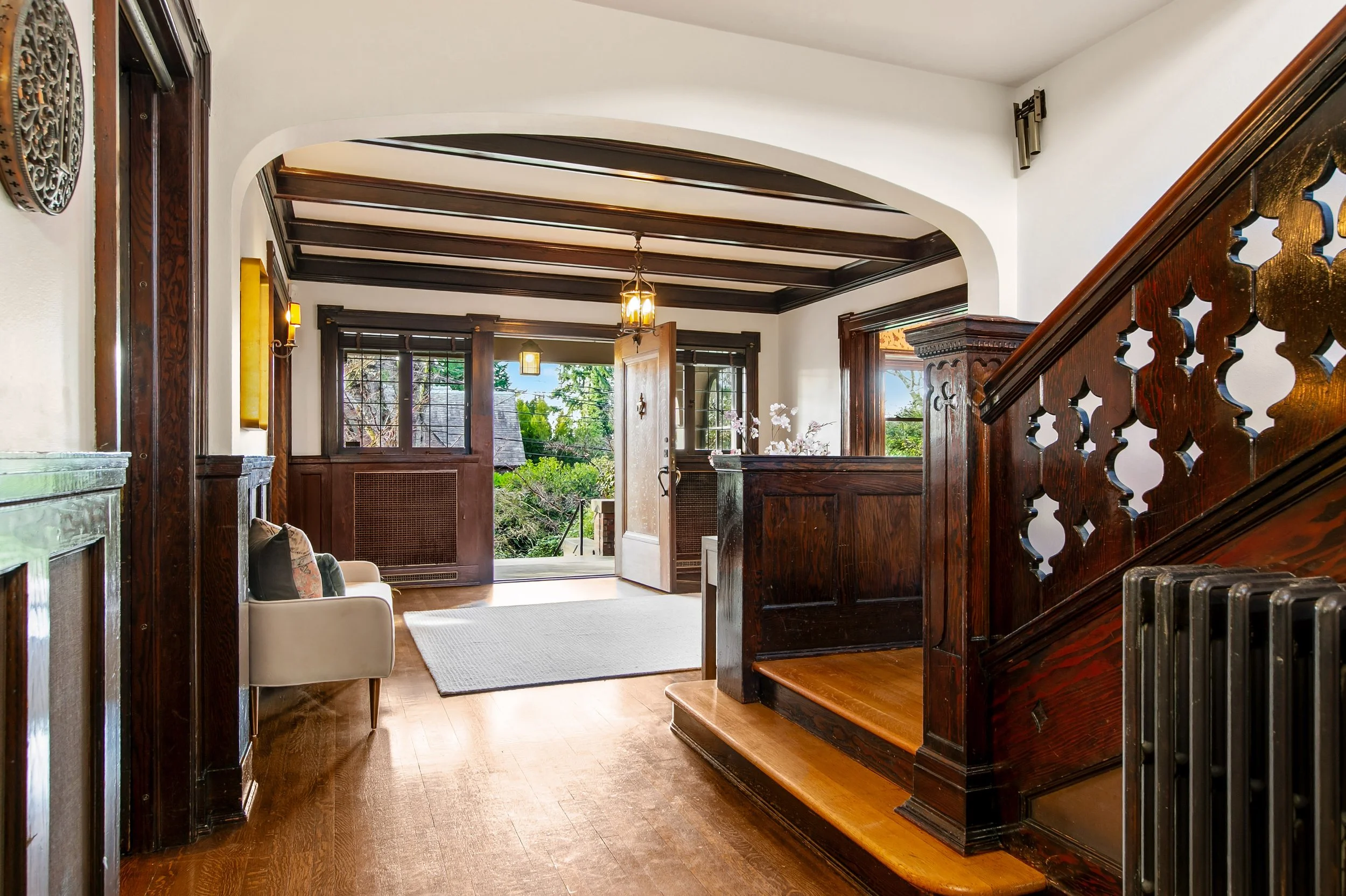 Foyer of a house with wooden stairs, dark wood trim, a white armchair with pillows, a runner rug, and an open front door leading outside