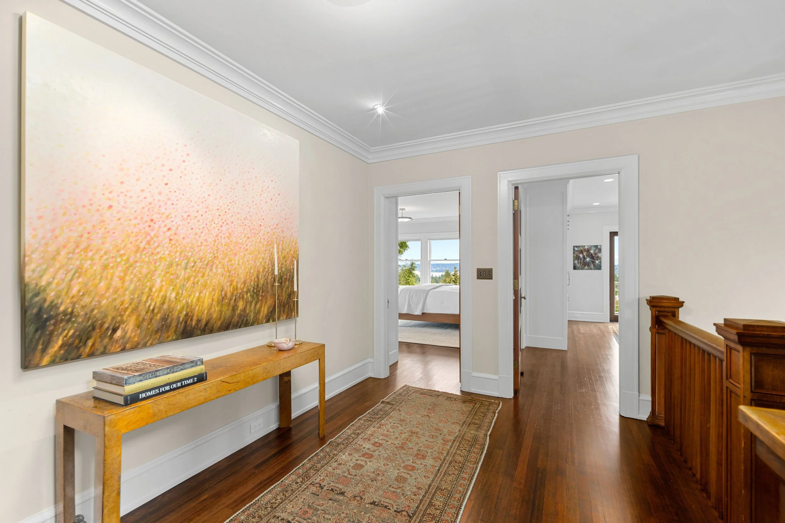 Hallway with wooden flooring, a large painting of a field, and a wooden console table with books and candles