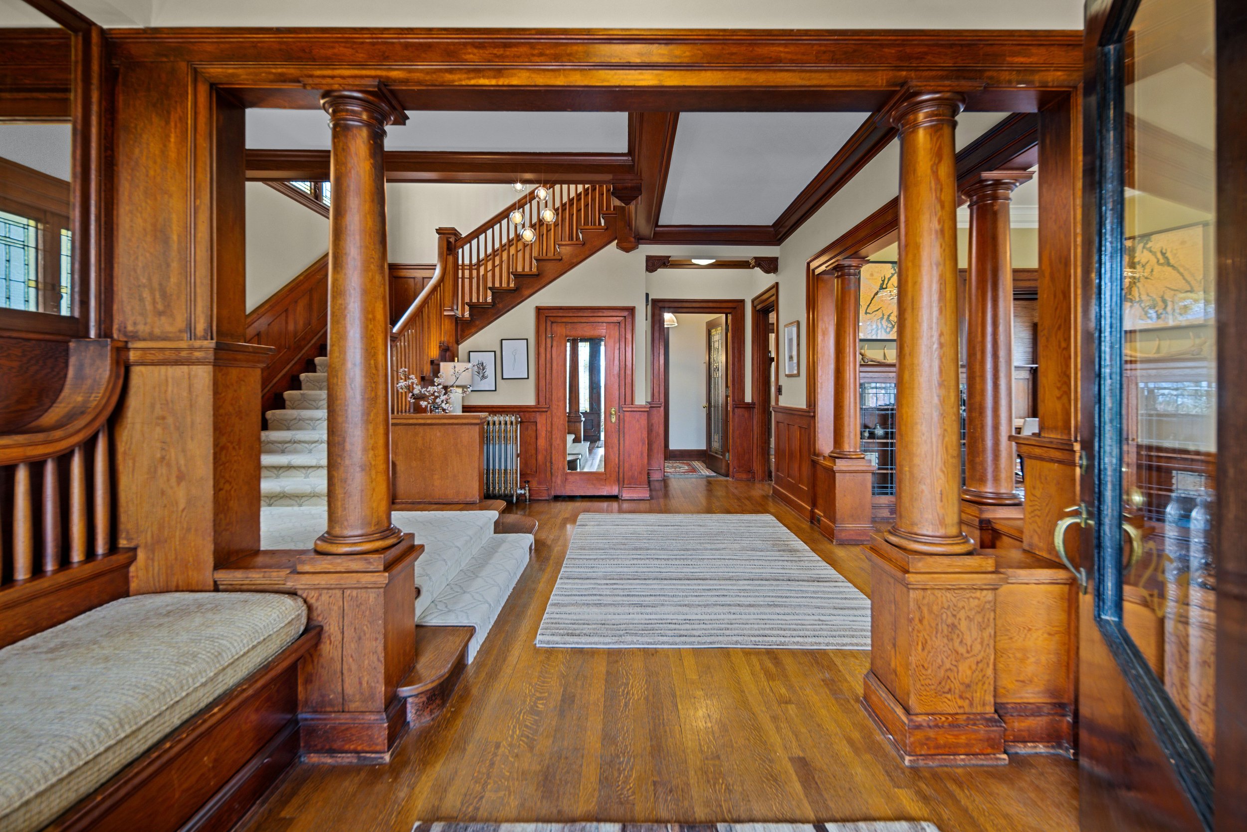 A spacious hallway in a house with wooden floors, walls, and trim, featuring a staircase with a wooden railing and a white carpeted runner. There are decorative framed pictures on the walls and a rug on the floor.