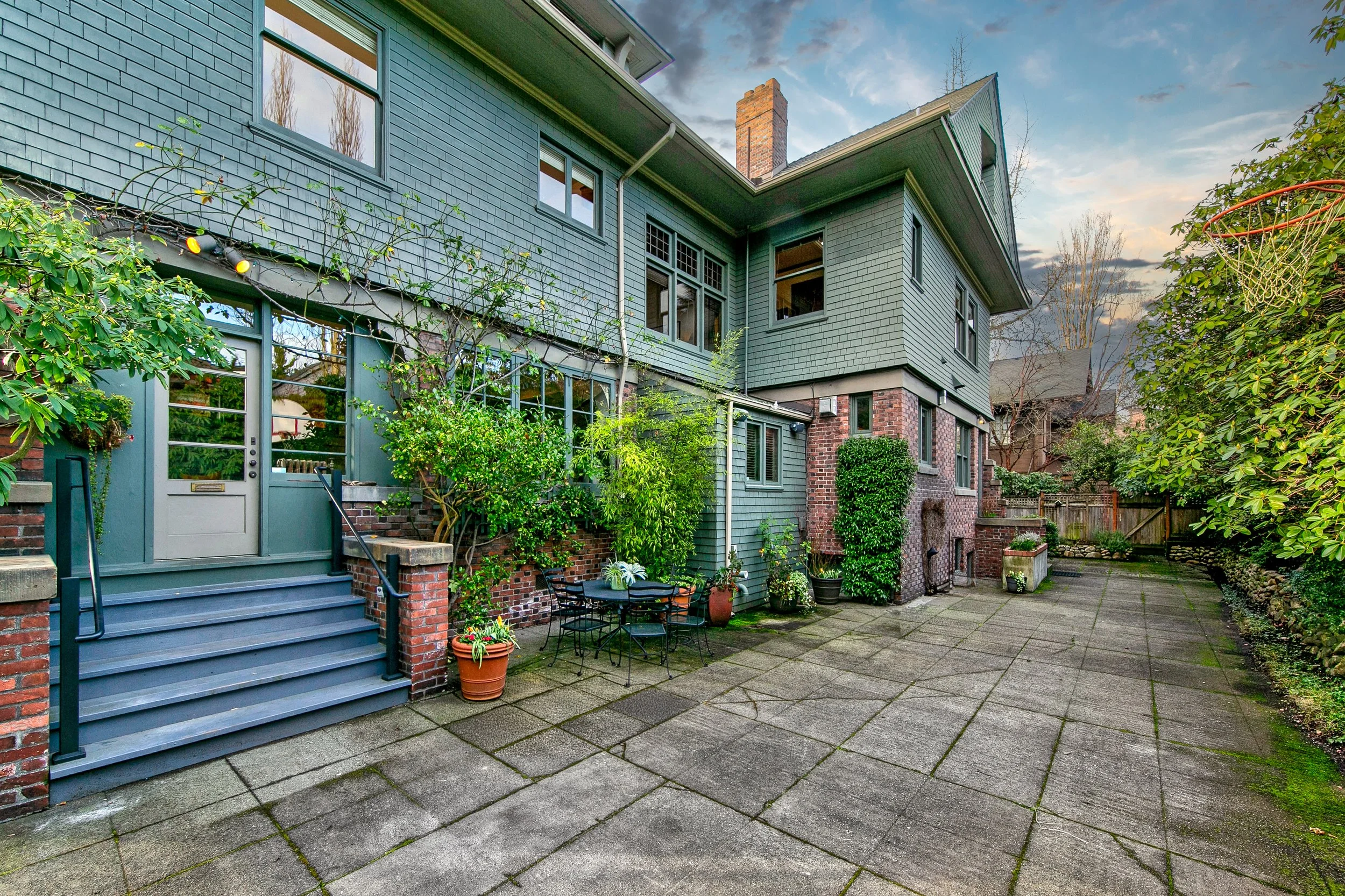 Backyard of a large green house with steps leading up to a door, patio furniture, potted plants, and a basketball hoop on the right, with a cloudy sky overhead.