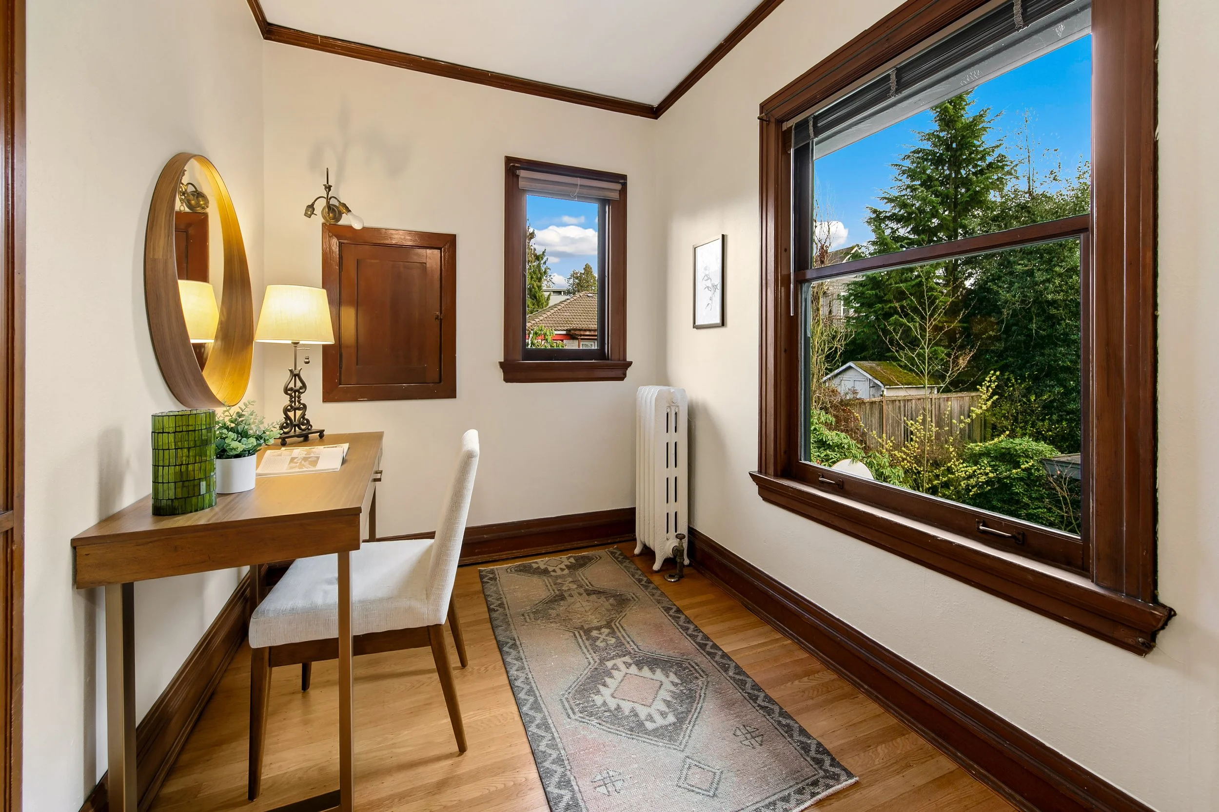 A small room with a wooden desk, a beige chair, a mirror, a table lamp, and decorative items. There are two windows with views of greenery and a blue sky, and a small radiator near the corner. A patterned rug lies on the wooden floor.