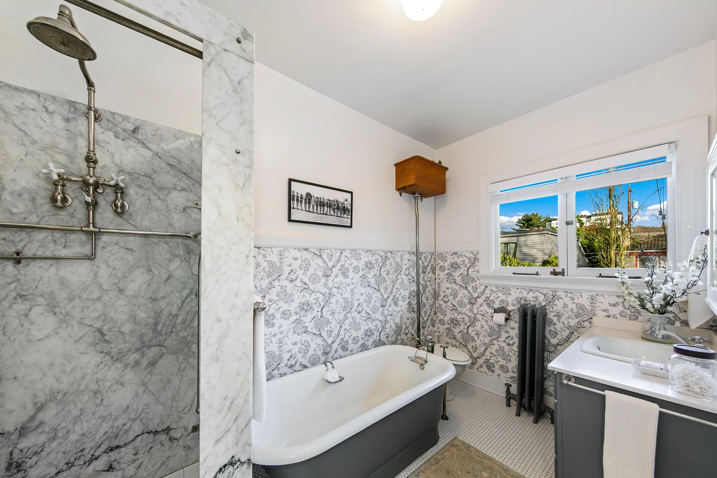 A vintage bathroom with a clawfoot bathtub, a shower area with marble walls, a sink with a dark cabinet, a window with a view of the outdoors, floral wallpaper, and various bathroom accessories.