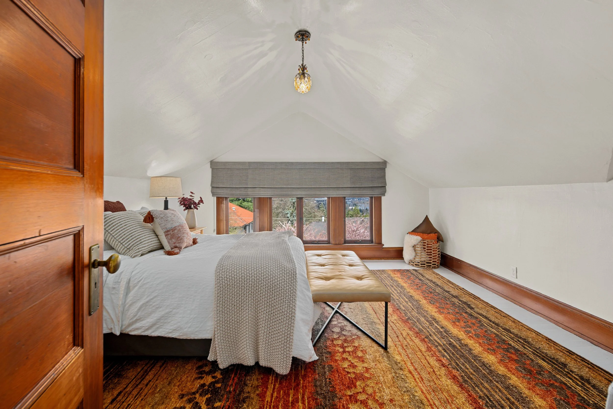 A cozy bedroom with a sloped ceiling. Features a large bed with multiple pillows, a side table with a lamp, a tufted bench at the foot of the bed, a window with a gray valance, and a colorful area rug on the floor.