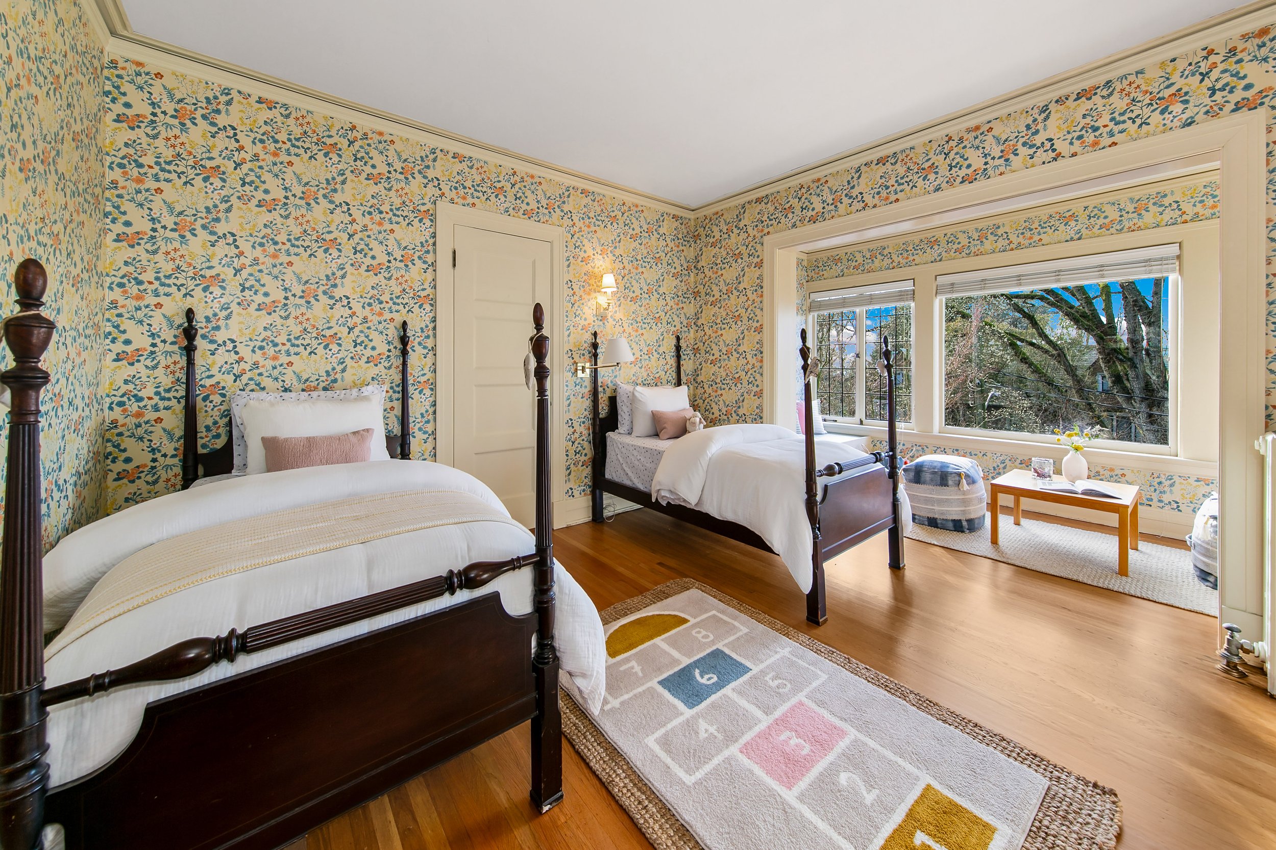 A cozy bedroom with two warm wood four-poster beds, floral wallpaper, hardwood floors, a large window showing trees, a small table near the window with a flower vase and books, and a hopscotch rug on the floor.