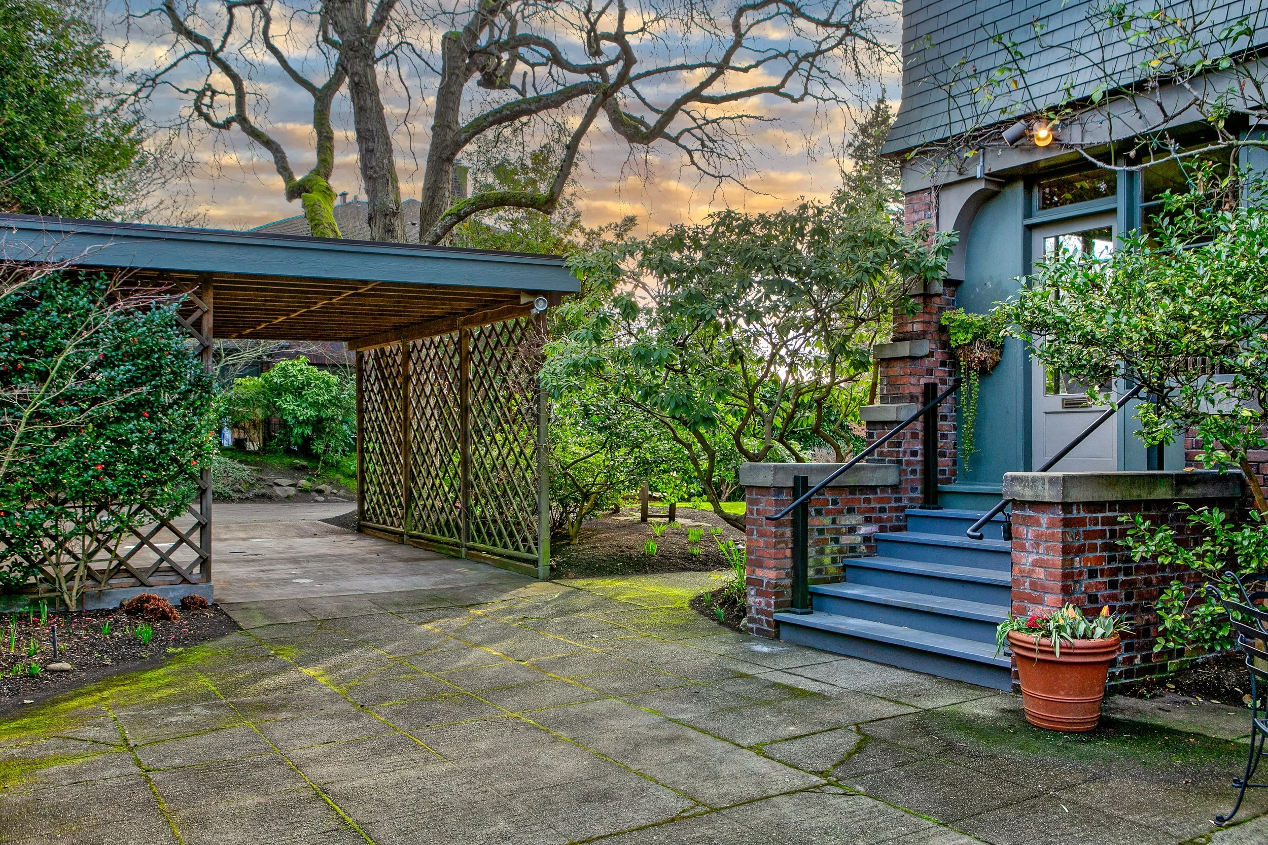 A backyard patio area with a staircase leading to a door on a brick house, surrounded by green plants and trees. There is a covered structure on the left side and potted plants near the stairs. The sky is partly cloudy at sunset.