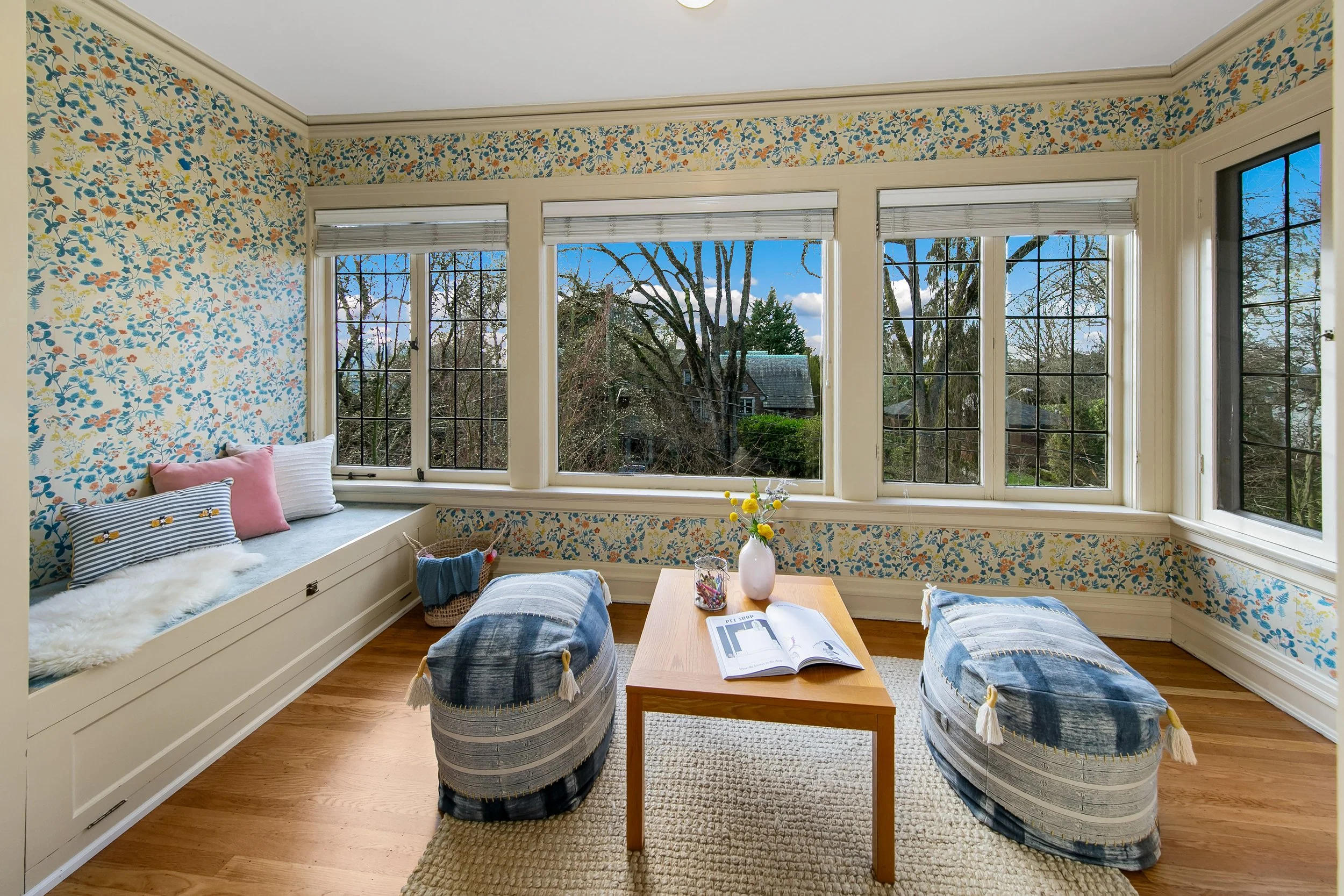 Sunroom with floral wallpaper, large windows, built-in window seat with cushions, two patterned poufs, a wooden coffee table with a book and flower vase, and hardwood floors.