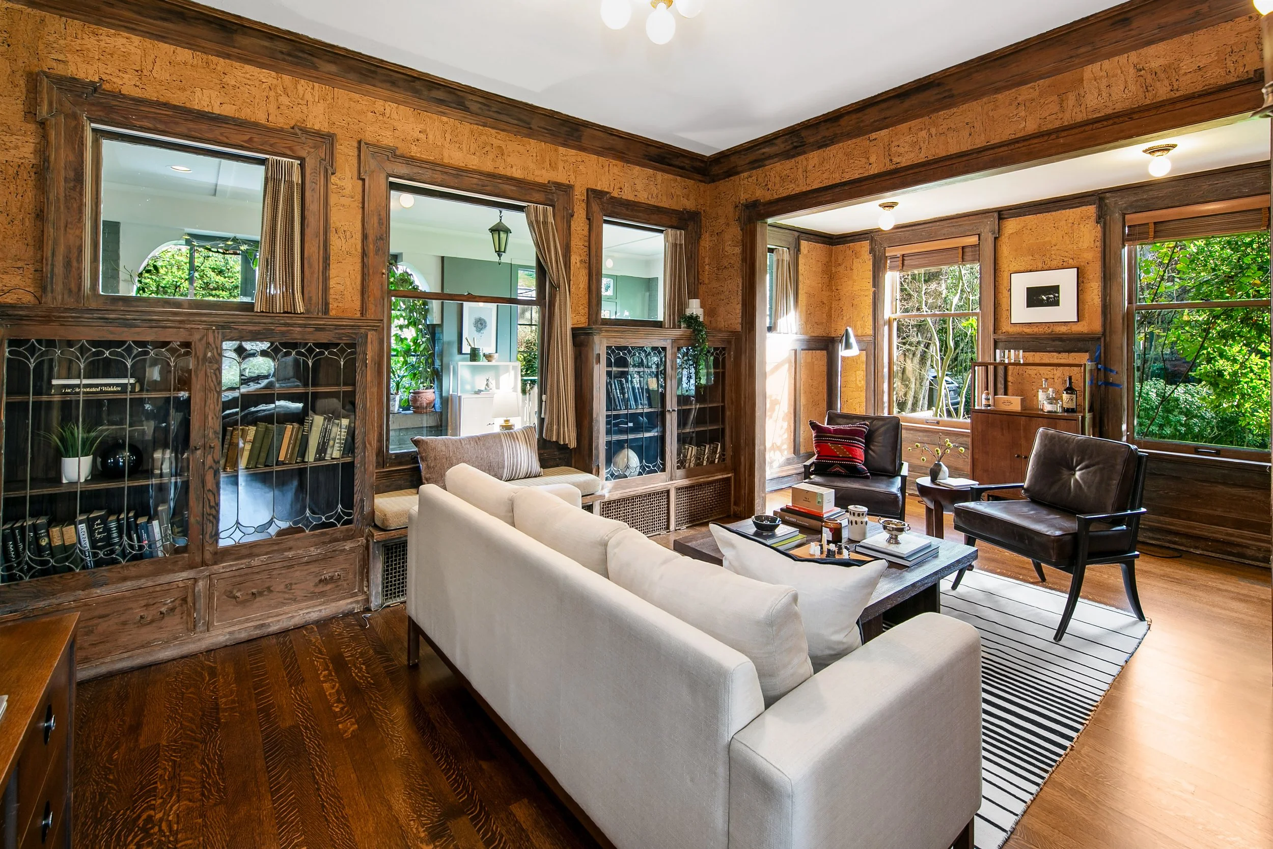 Living room with wooden walls, large windows, and vintage furniture including a white sofa, leather armchairs, bookshelves, and a coffee table, with greenery outside the windows.